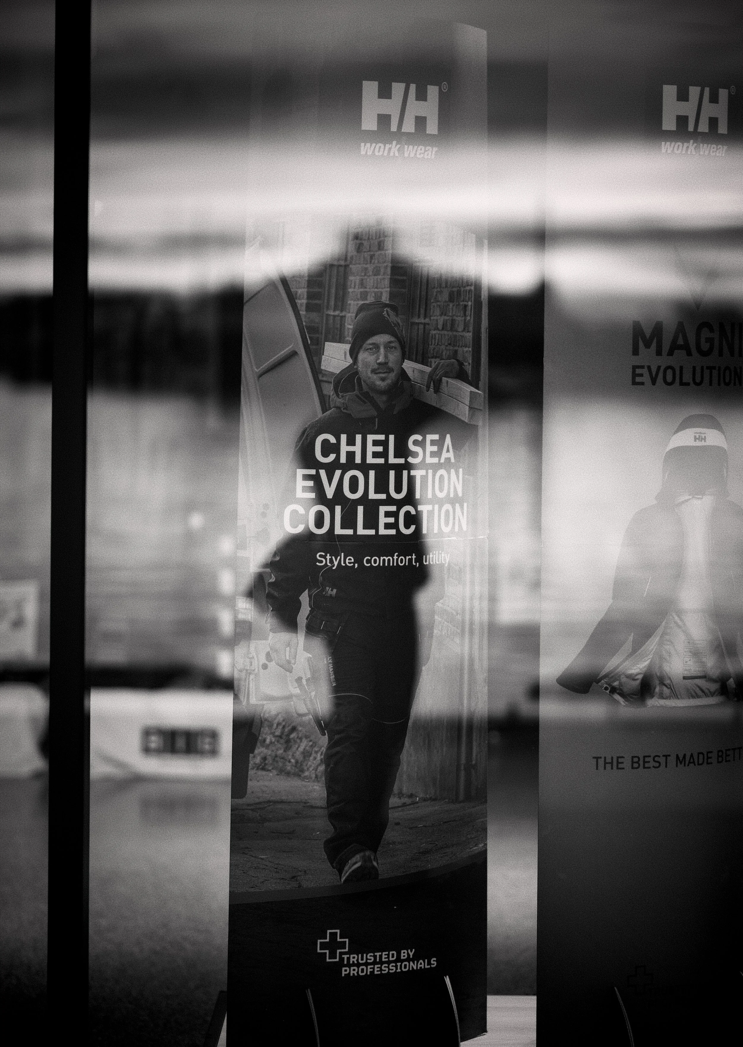 Black and white advertisement poster for Chelsa Evolution Collection showing a man holding a power drill, walking outdoors in front of a brick building.