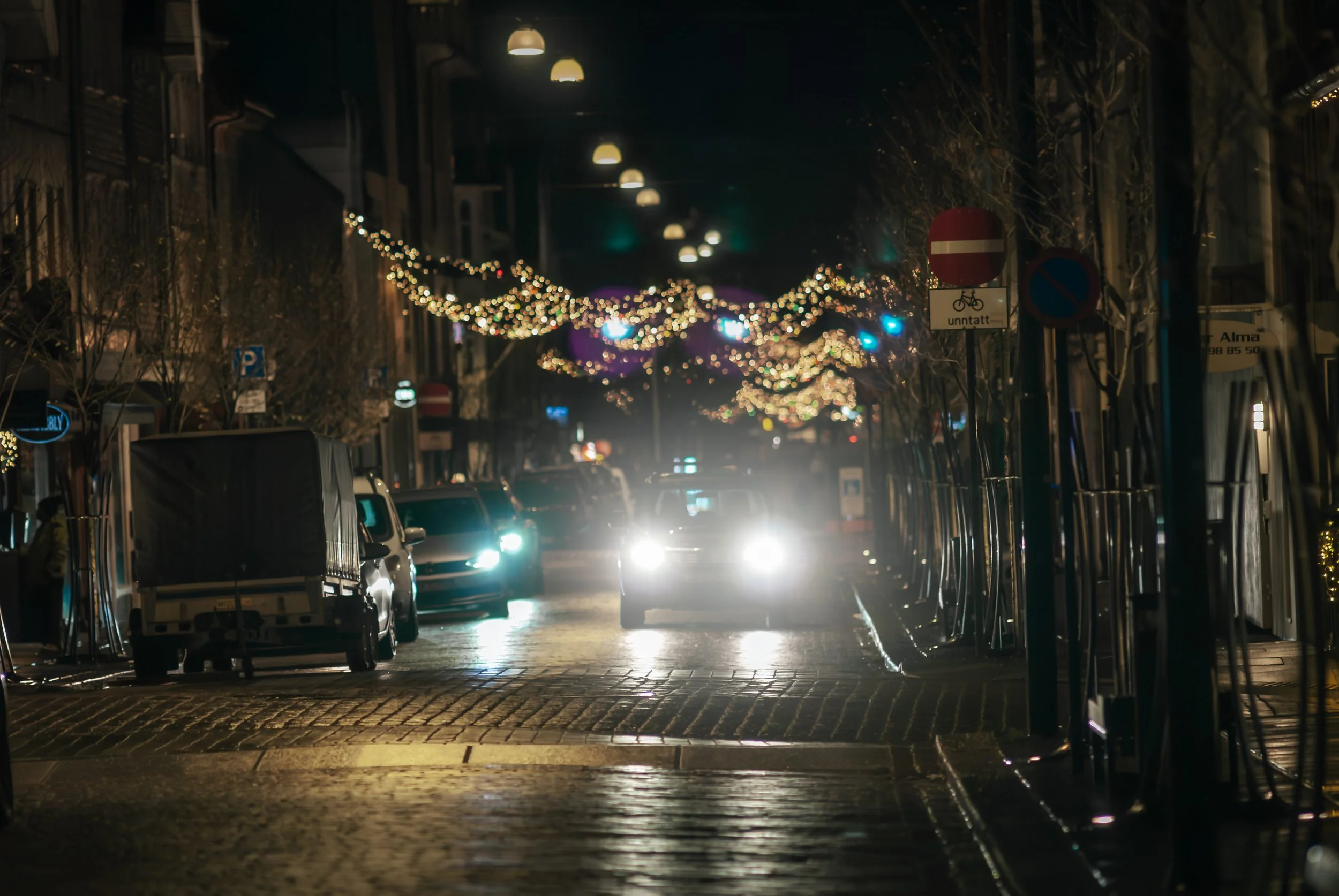 Nighttime city street decorated with festive string lights, parked cars, and street signs, with a few shining car headlights.