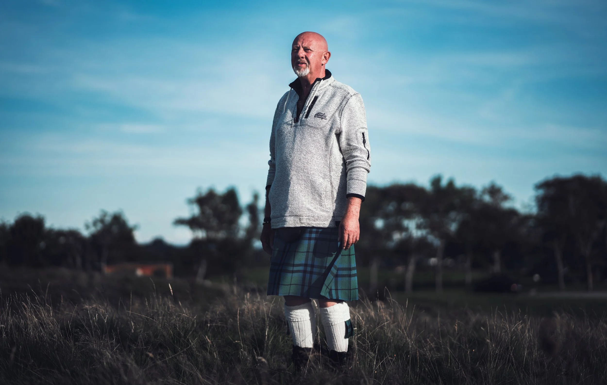 En eldre mann står i et åpent landskap under en blå himmel, iført en lys genser, kilt, og hvite lagsko.