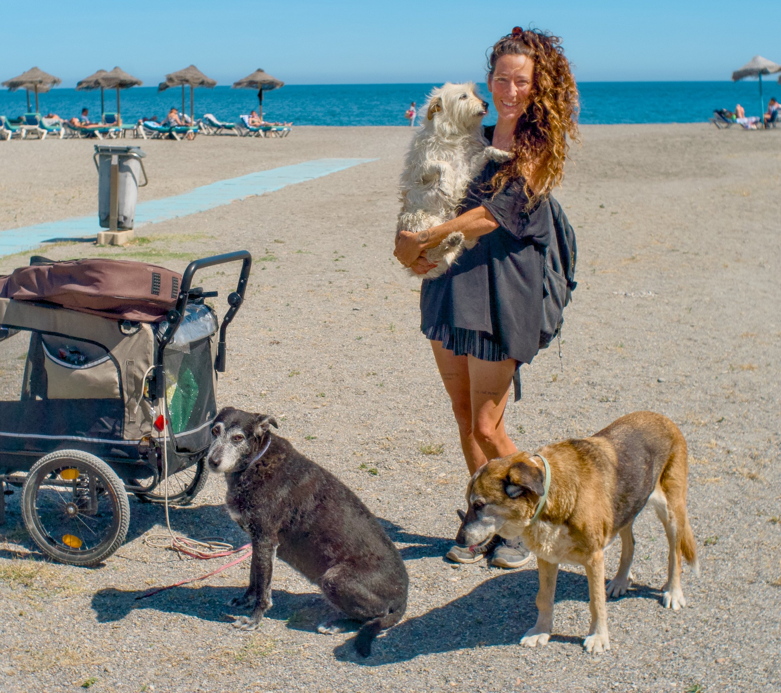 En kvinne med krøllete hår smiler mens hun holder en liten hvit hund på fanget på en strand, med flere mennesker som soler seg under parasoller i bakgrunnen.