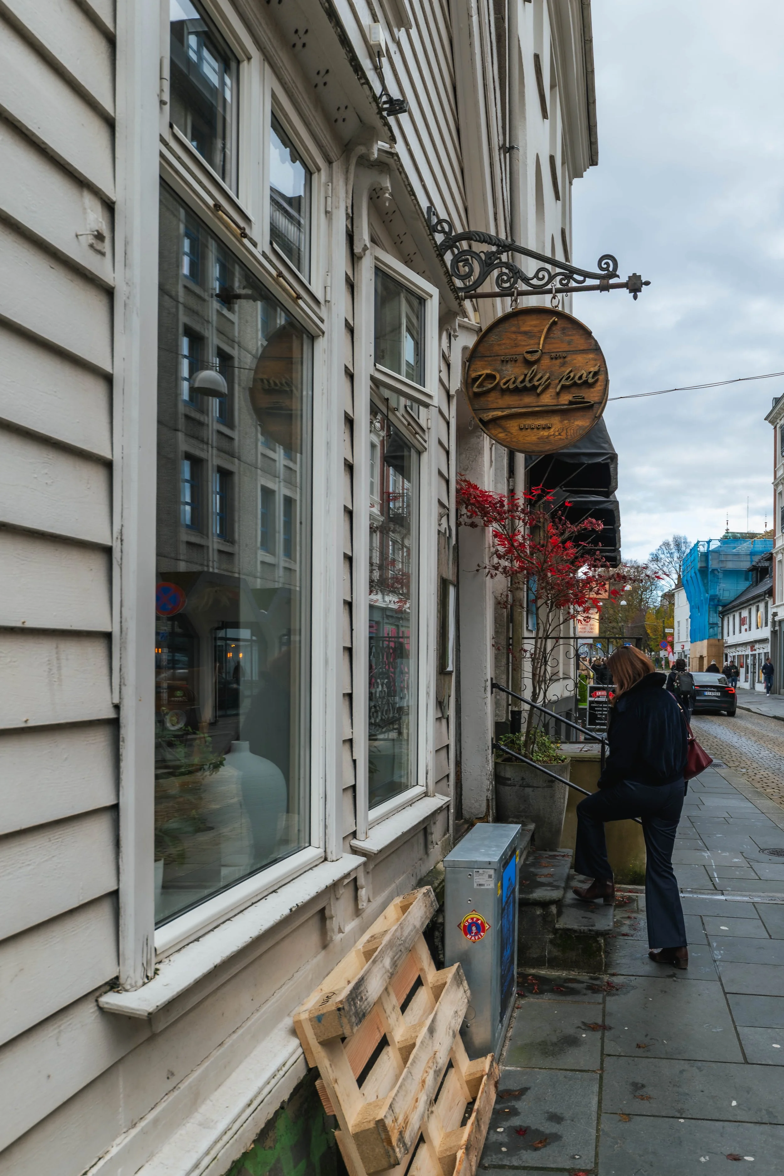 A street view showing a woman climbing the steps outside a shop with a wooden sign that reads 'Daily Pot'. The building has beige siding and large windows, and there are some red leaves on a small tree near the entrance. The sky is cloudy.