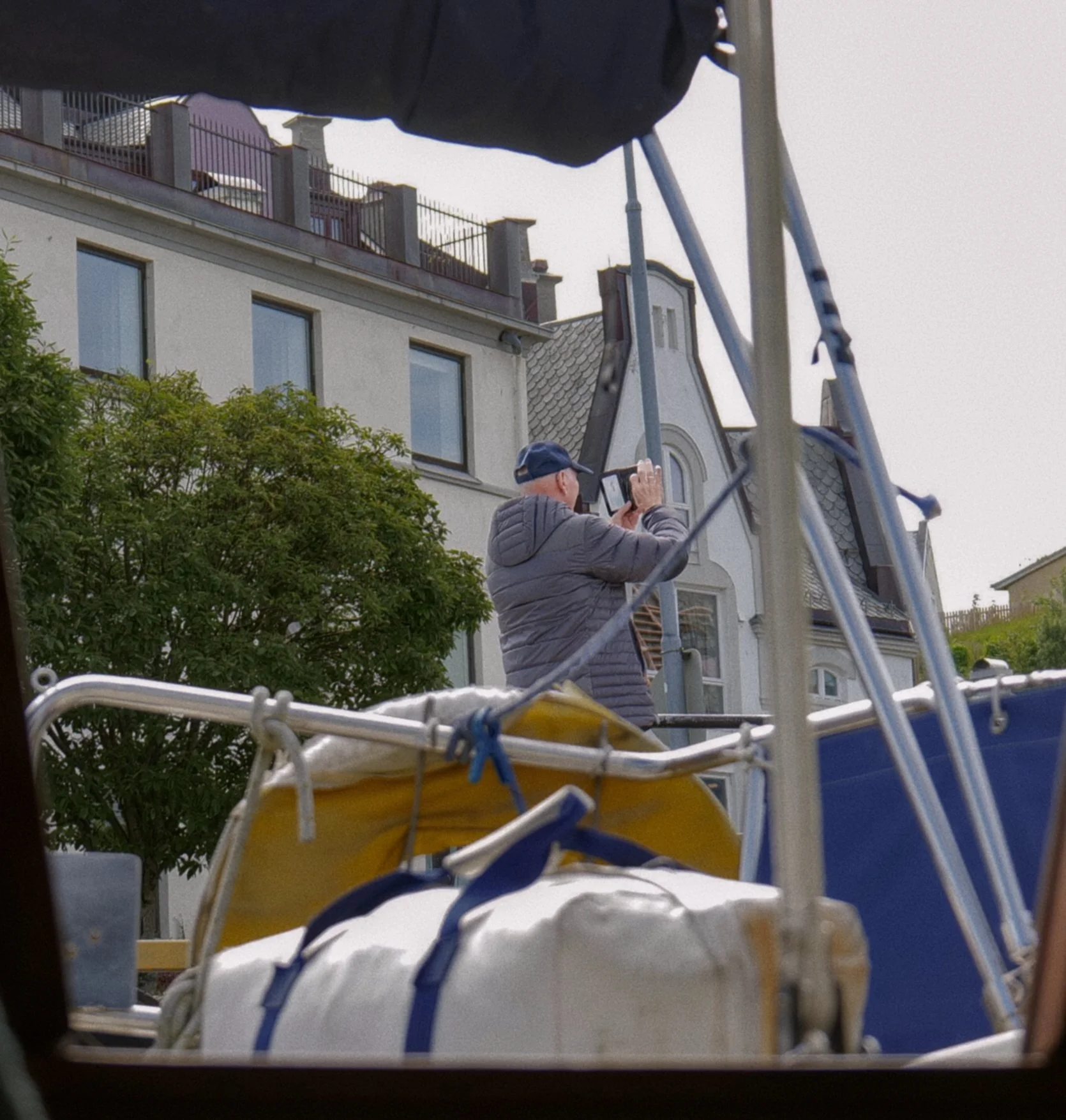 A man with a blue cap taking photos with a phone from a boat or dock, with a residential building behind him and various equipment and supplies in the foreground.