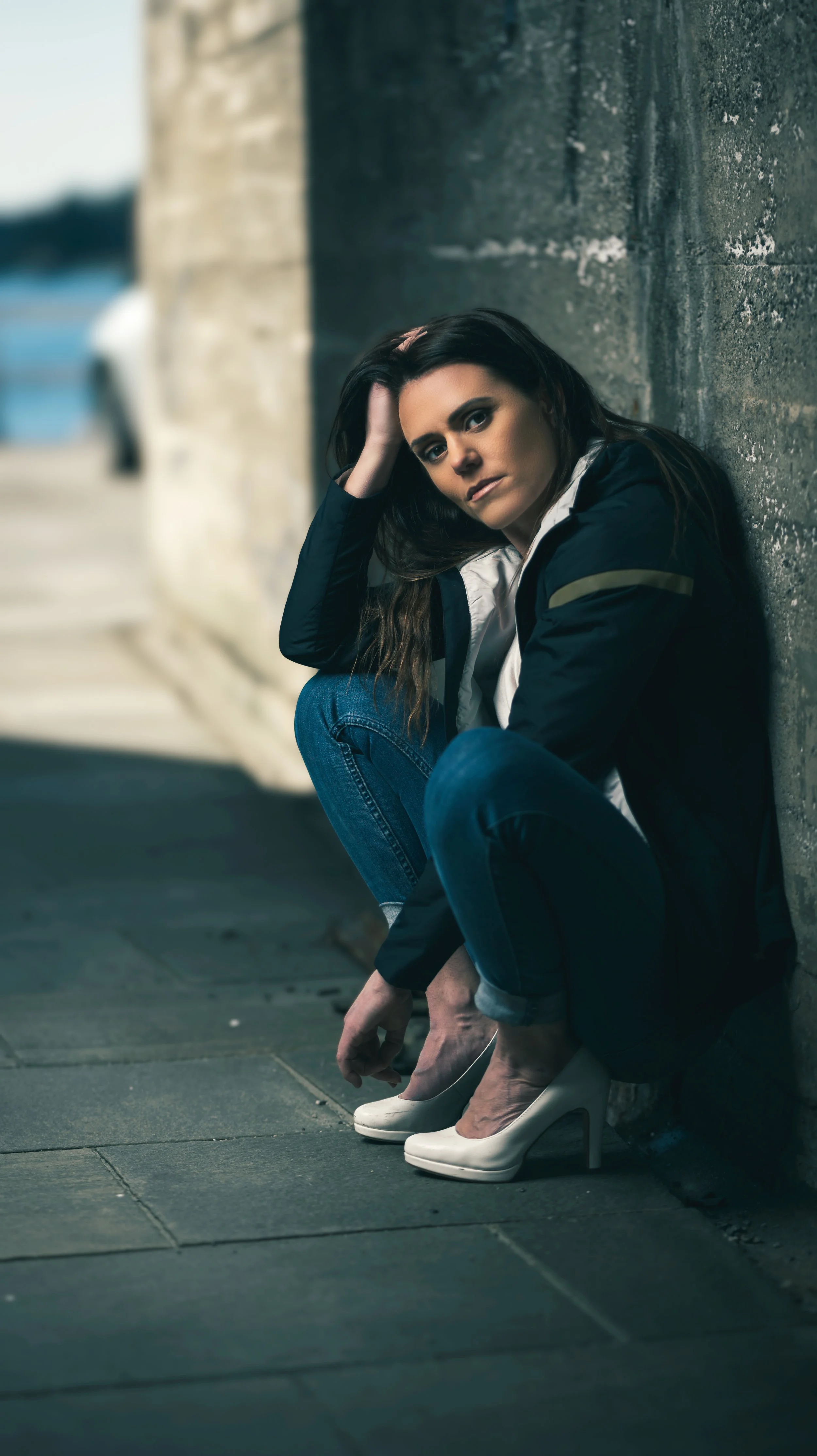 A woman with long dark hair sitting against a stone wall, wearing blue jeans, a black jacket, and white high heels, looking at the camera with a serious expression.