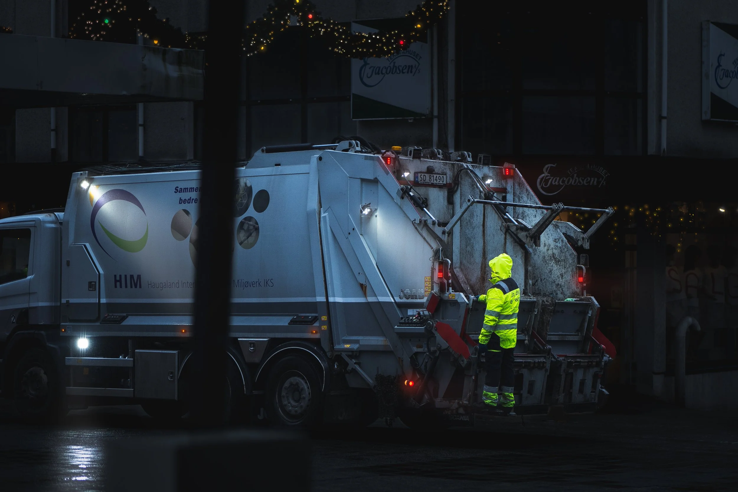A sanitation worker in a yellow high-visibility jacket and pants standing beside a street sweeper at night. The street sweeper is white with some logos and text, and it is cleaning the street. The background shows some lit decorations, possibly holid