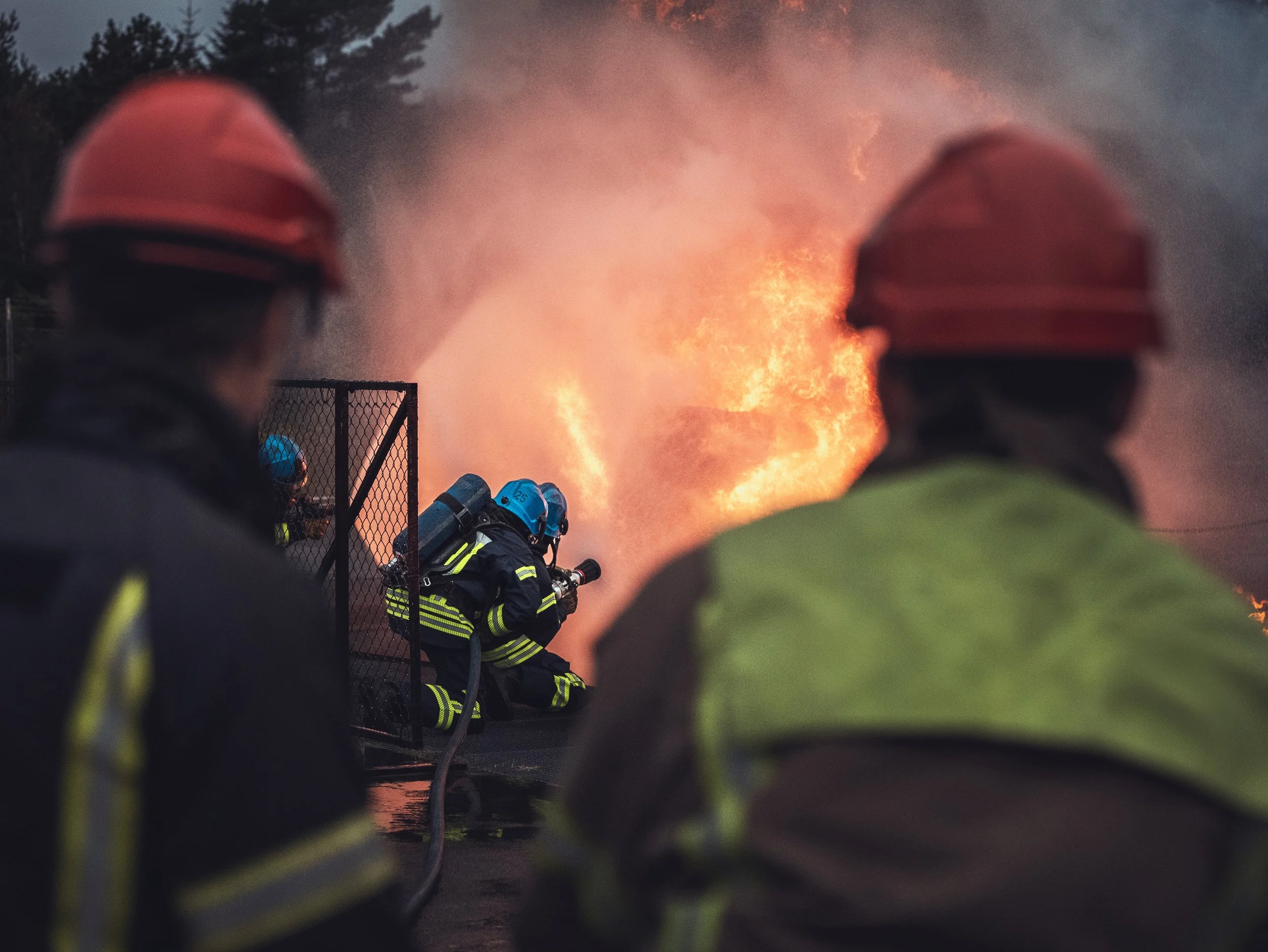 Firefighters in protective gear and helmets working to control a large fire seen from behind a chain-link fence, with flames and thick smoke in the background.