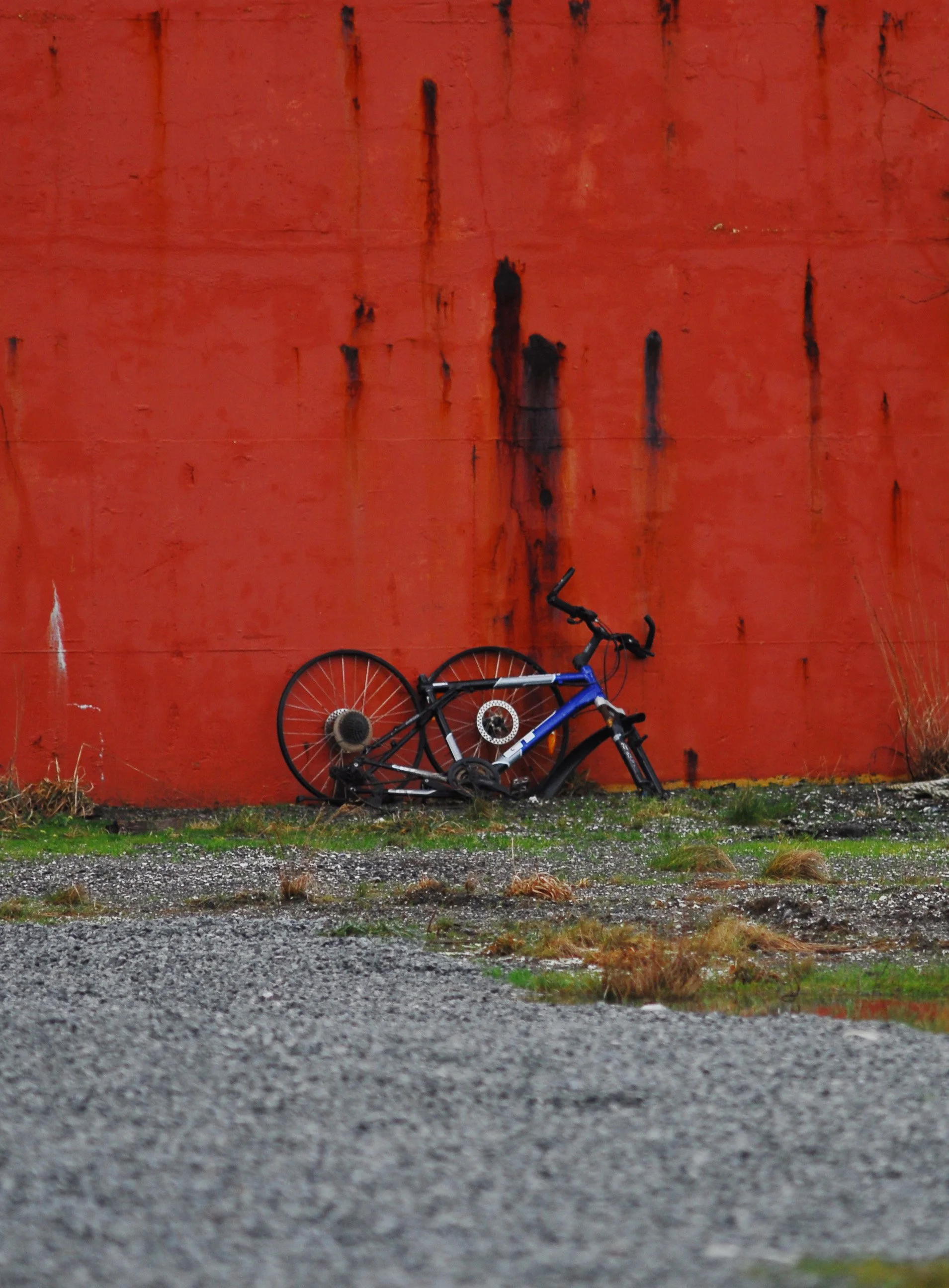 A broken bicycle with the front wheel and part of the frame on the ground, leaning against a large, red, rusty wall with black stains.