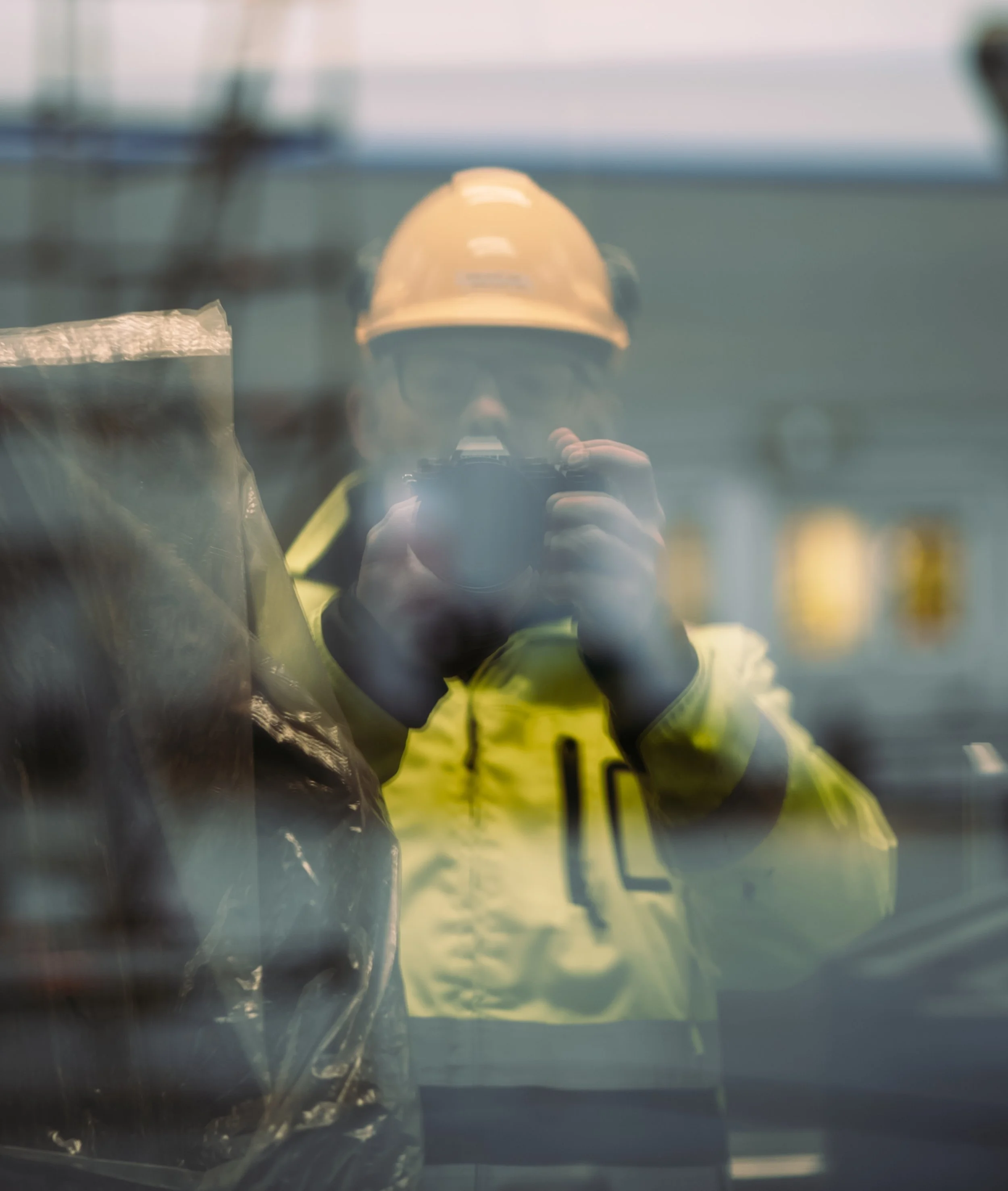 Person wearing safety helmet and reflective jacket taking a photo through a reflective surface.