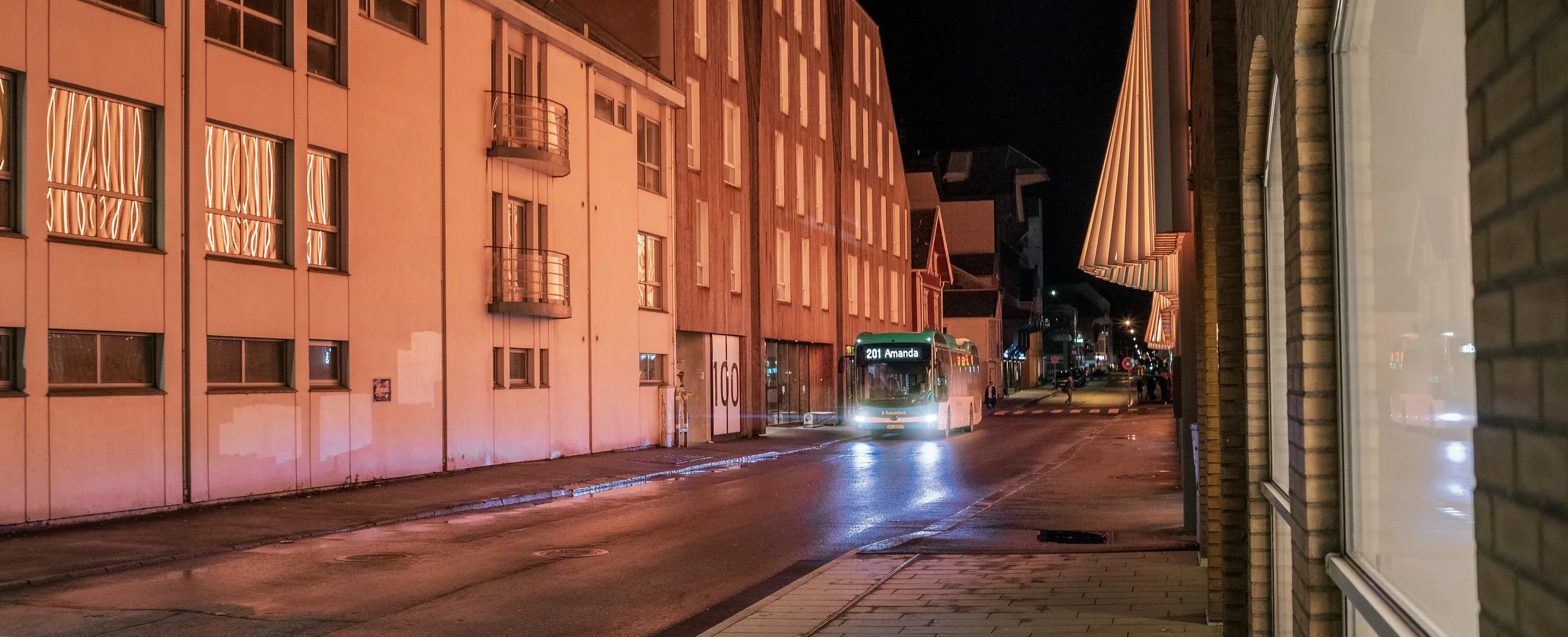 A city street at night with a bus labeled 201 Amanda approaching the curb. The street is wet and illuminated by streetlights. Buildings with awnings and windows line both sides of the street.