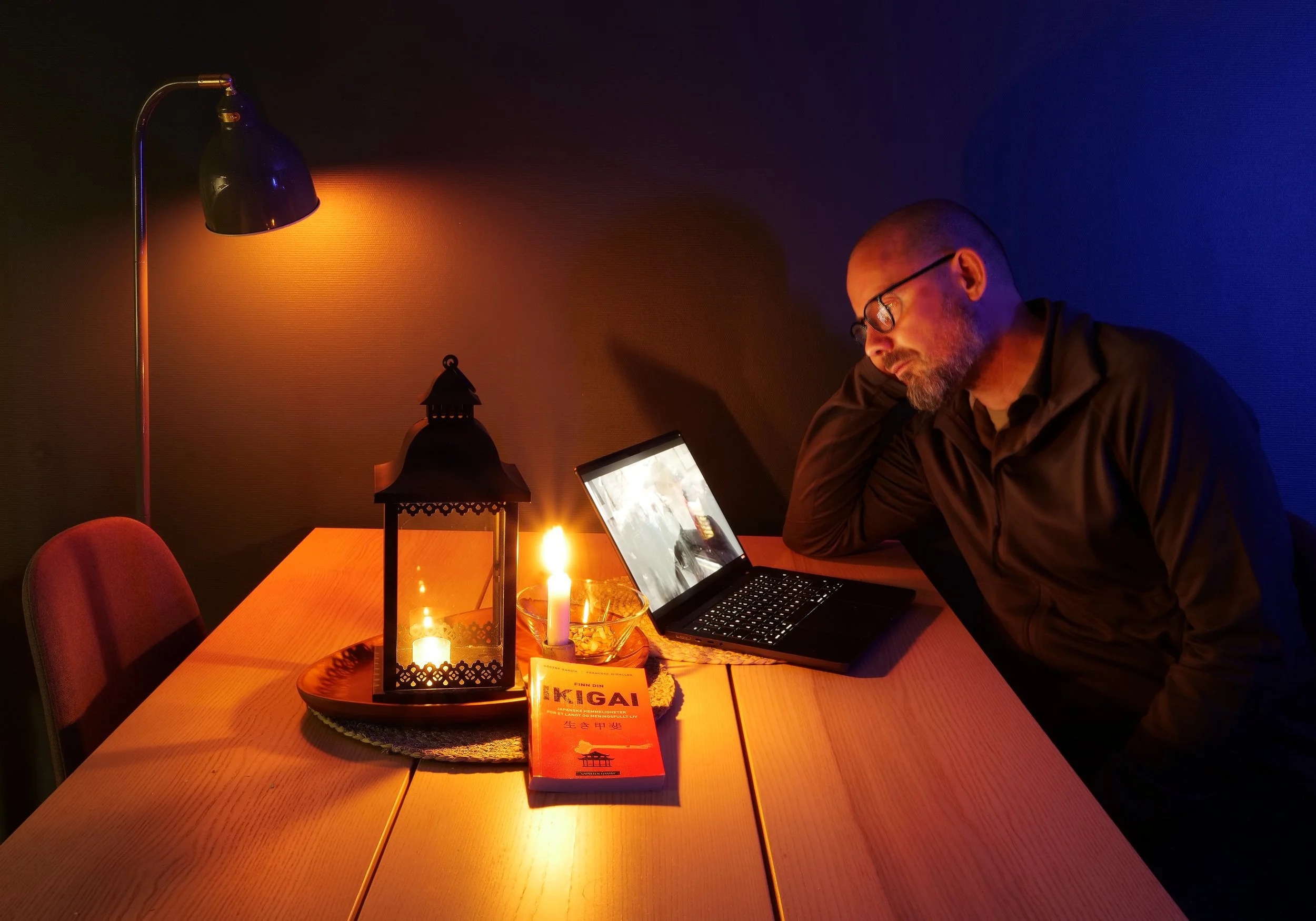 A man sitting at a wooden dining table with a laptop, a candle, a lantern, and a book, in a dimly lit room with warm and cool lighting.