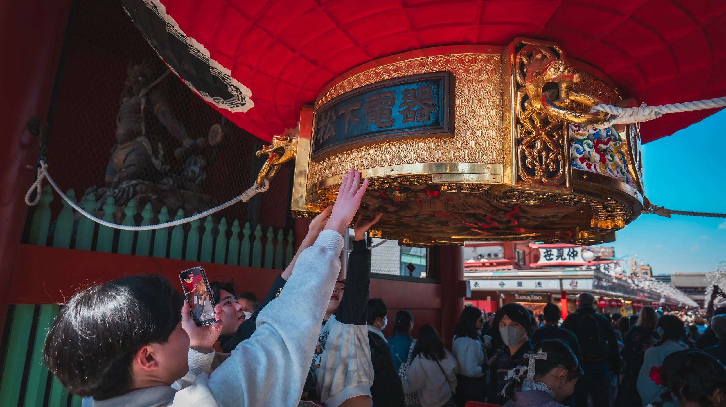 People adjusting and preparing a large, ornate Japanese lantern on a temple or shrine. The lantern is gold, with traditional Japanese characters and decorative designs, surrounded by a crowd of visitors.