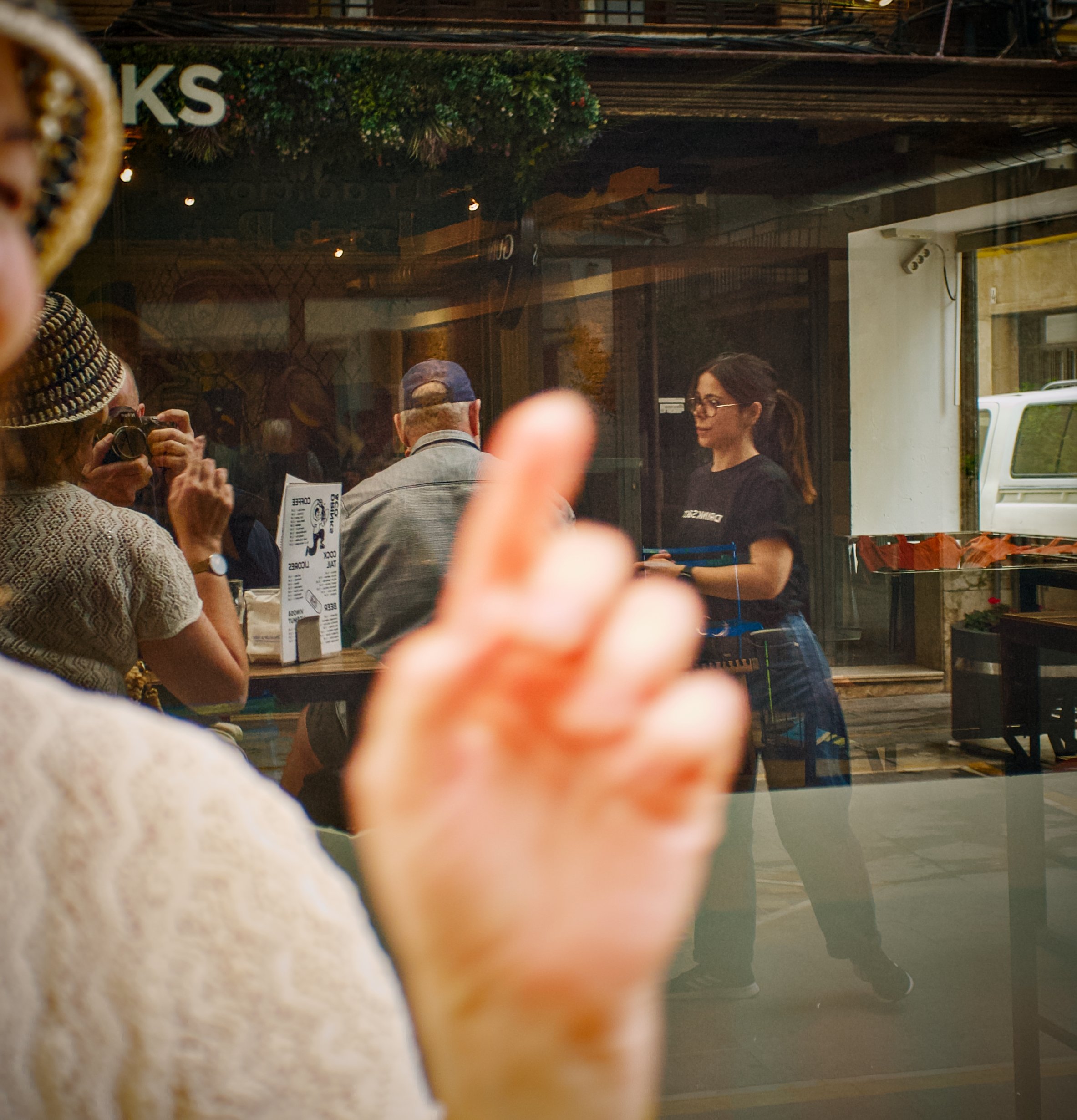Ukjent gruppe mennesker som snakker og tar bilder i en butikk eller kafé, sett gjennom et glassvindu, med en meny på bordet.