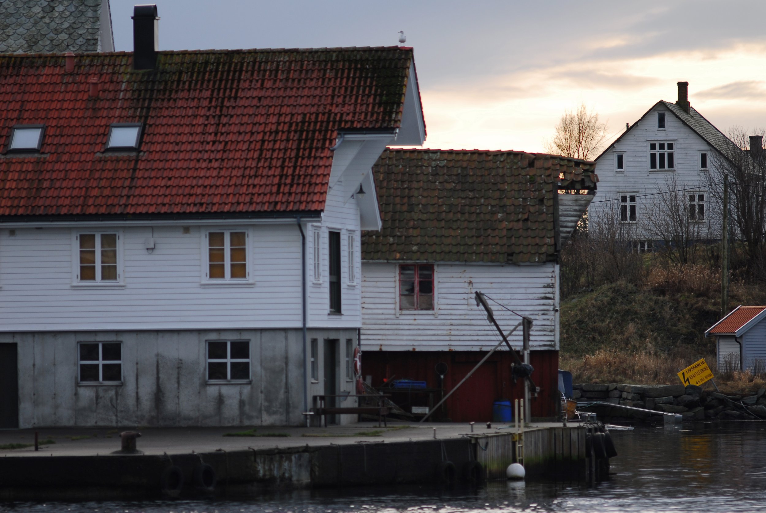 A riverside scene with a white wooden house and a smaller building, both with tiled roofs. The larger house has a concrete foundation and several windows. The smaller building shows signs of weathering and is partly obscured by a boat or dock. A yell