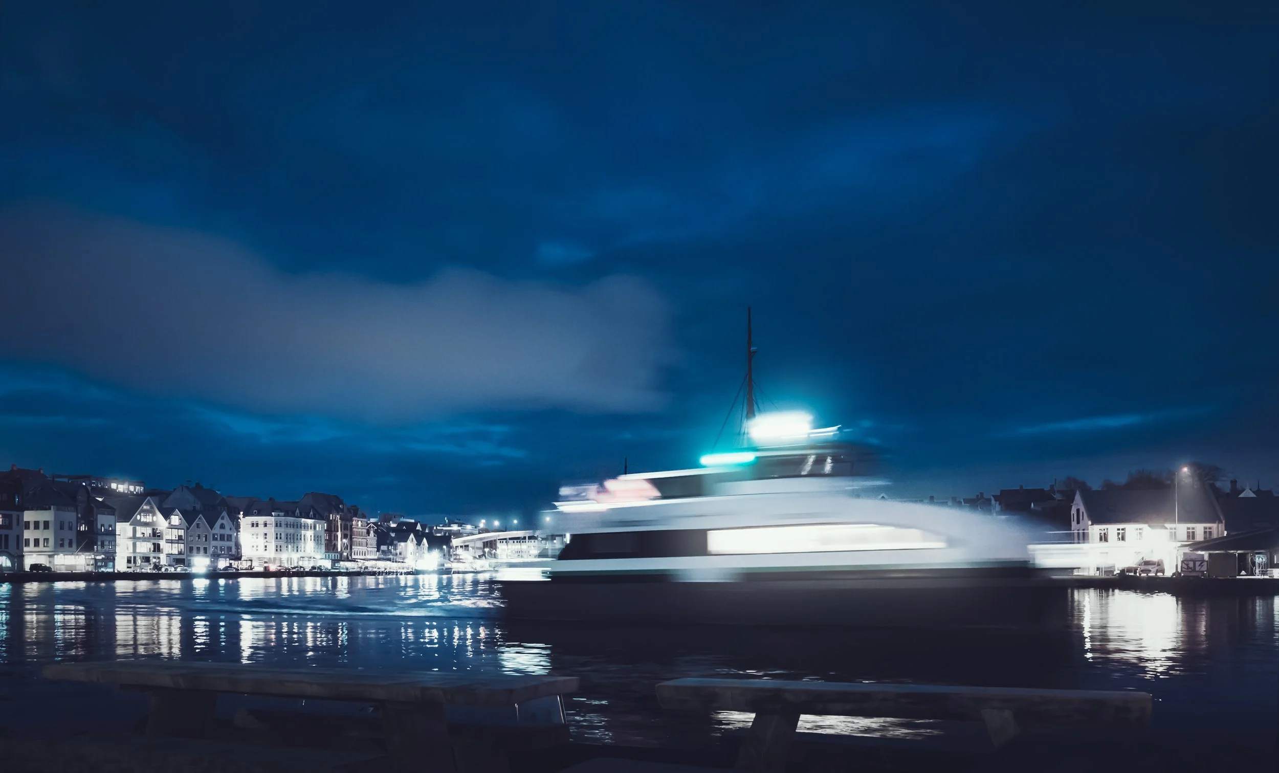 A yacht moving quickly through a harbor at night, creating a motion blur with city buildings illuminated in the background and dark cloudy skies above.