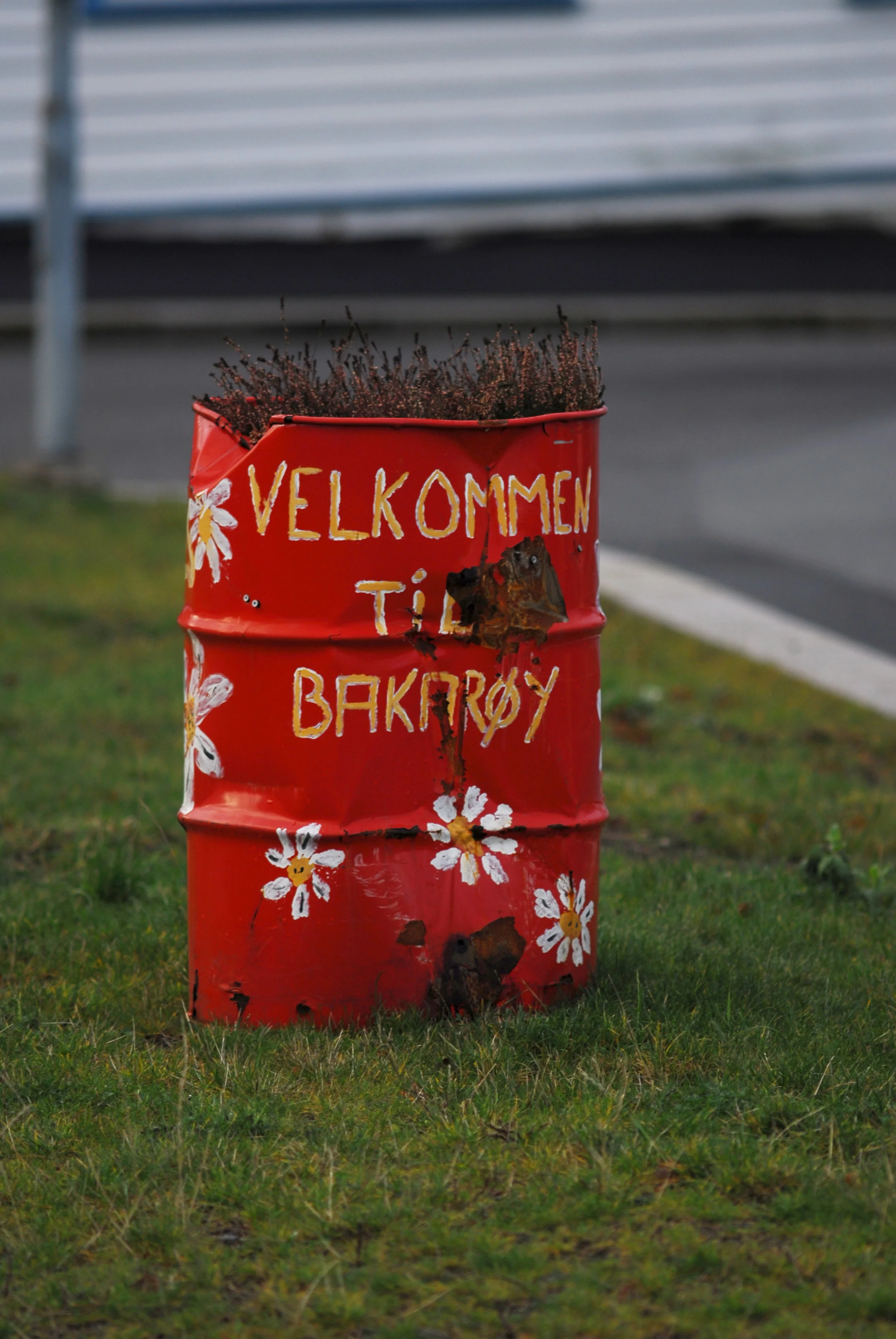 Red metal barrel with white daisy flower decorations and the words 'Velkommen til bakery' painted on it, placed on grass near a sidewalk.
