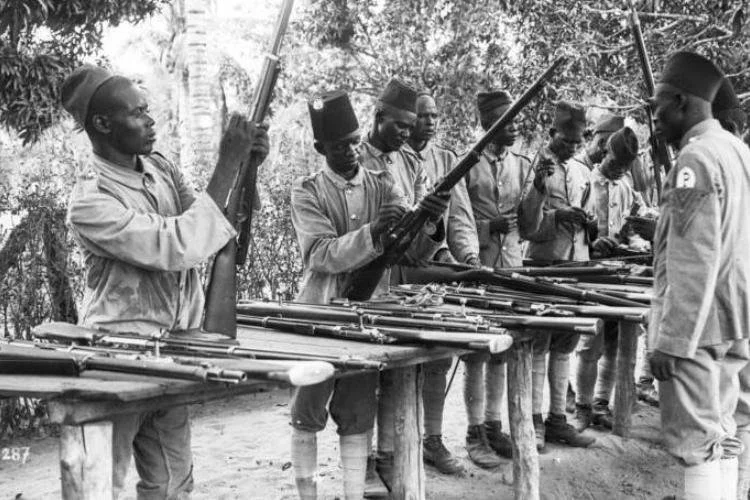 Black-and-white photograph of African police askaris cleaning rifles on wooden tables in German East Africa, circa 1906–1914.