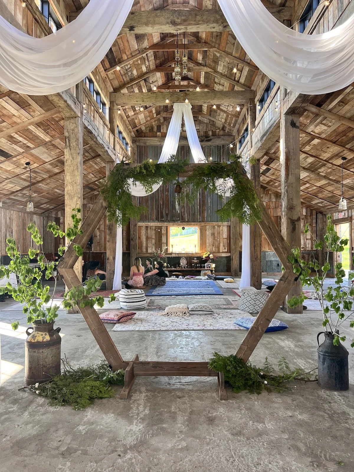 Interior of a rustic barn at a retreat, with draped white fabric and string lights hanging from the ceiling, a wooden diamond-shaped frame with greenery in the foreground, and people sitting on the floor with cushions and rugs in the background.