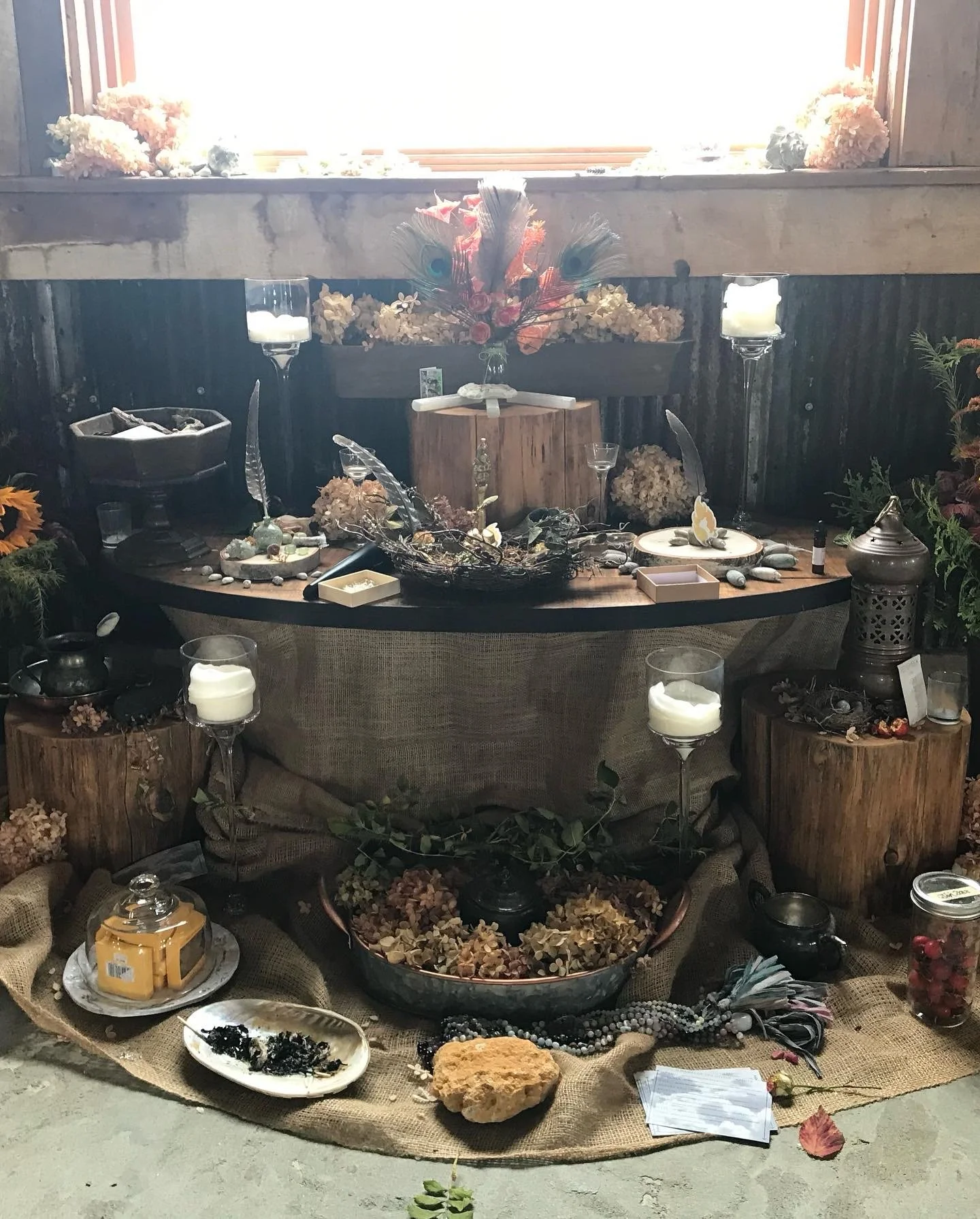 Decorative altar with flowers, candles, and various small objects on a burlap cloth, wooden stumps, and a black table.
