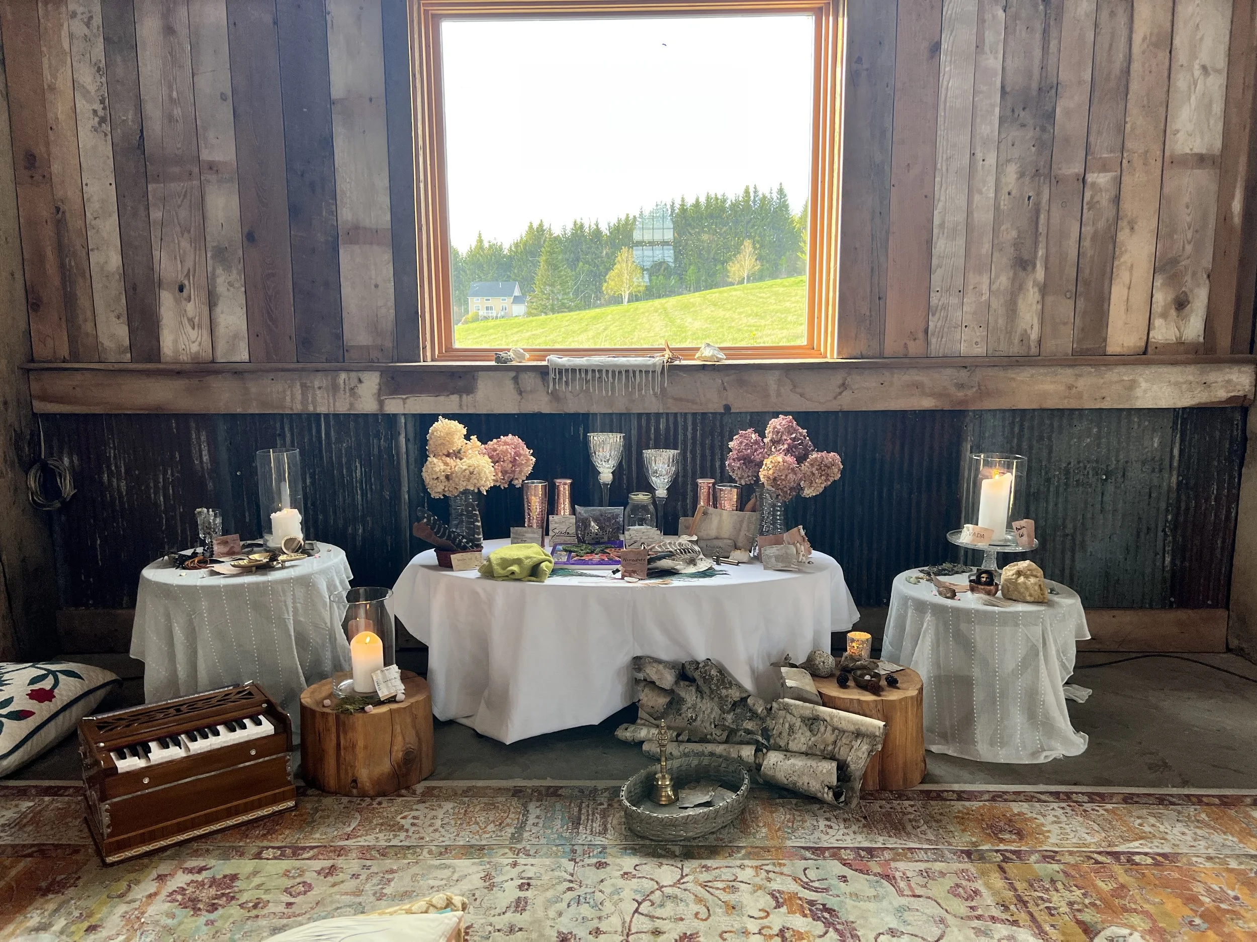 A rustic display of tables with white tablecloths holding candles, flowers, and decorative objects in front of a large window showing a green landscape outside. 