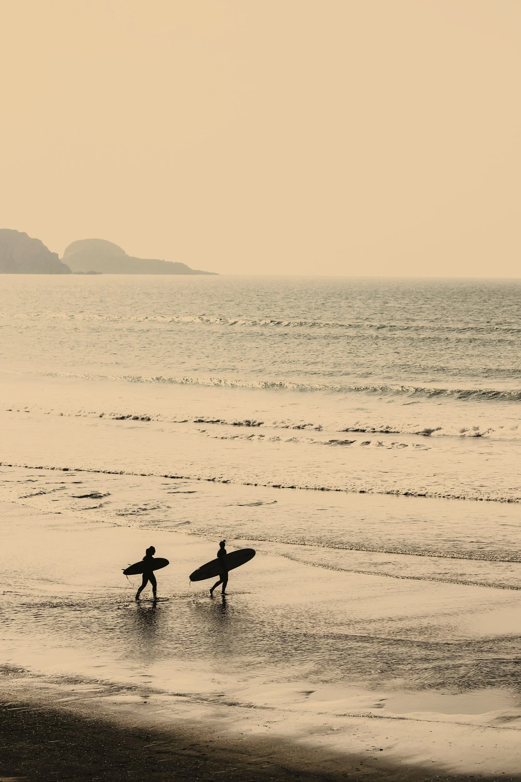 Two surfers walking out of the ocean carrying surfboards at sunset or sunrise on a beach.