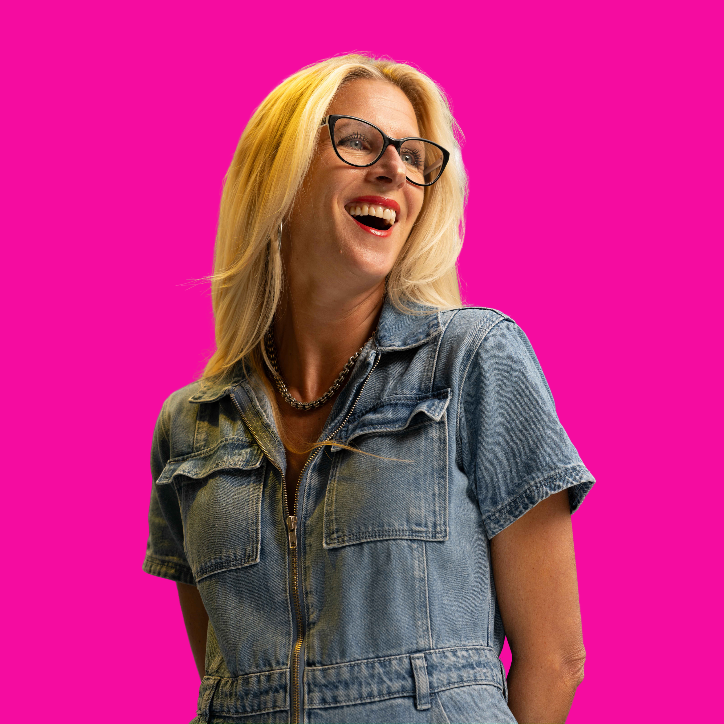 A woman with blonde hair and glasses smiling, wearing a denim jacket and necklace, against a bright pink background.