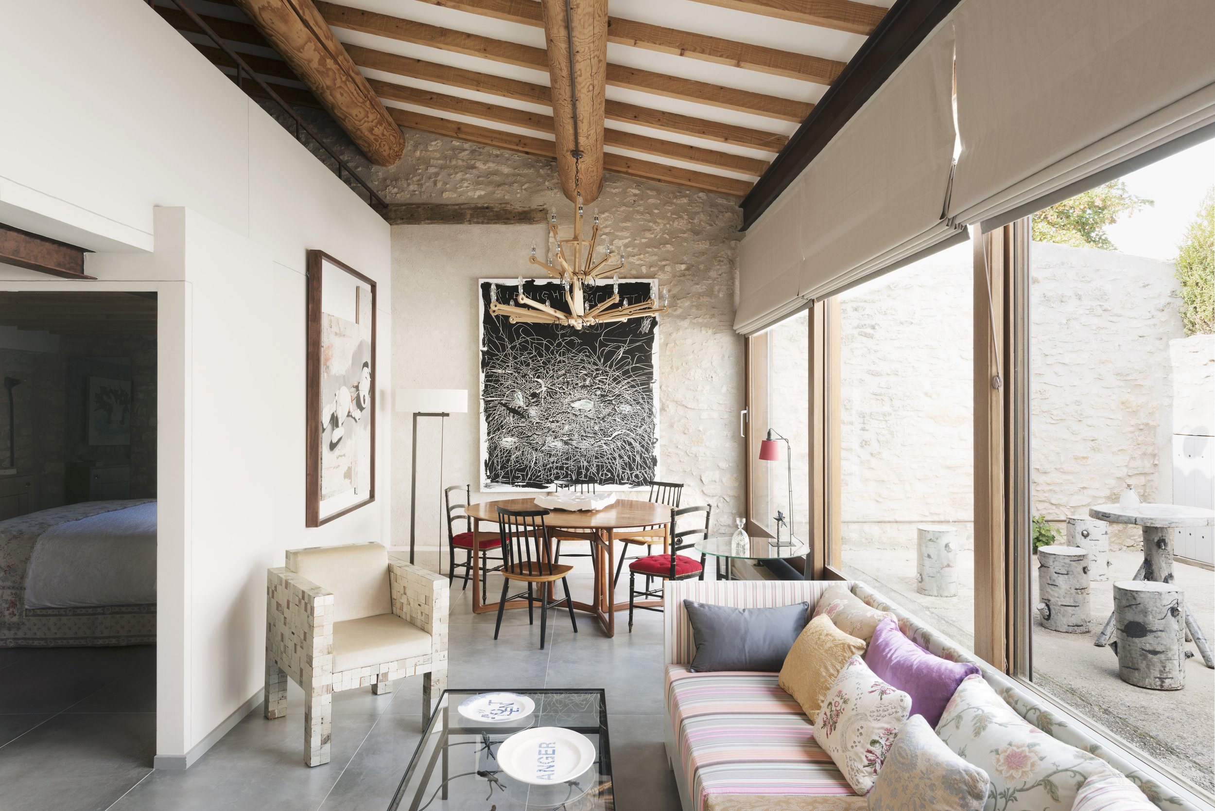 Interior of the open plan living and dining room of the restored stone barn, with exposed wooden beams and floor to ceiling glazing to one side, opening onto a walled courtyard.