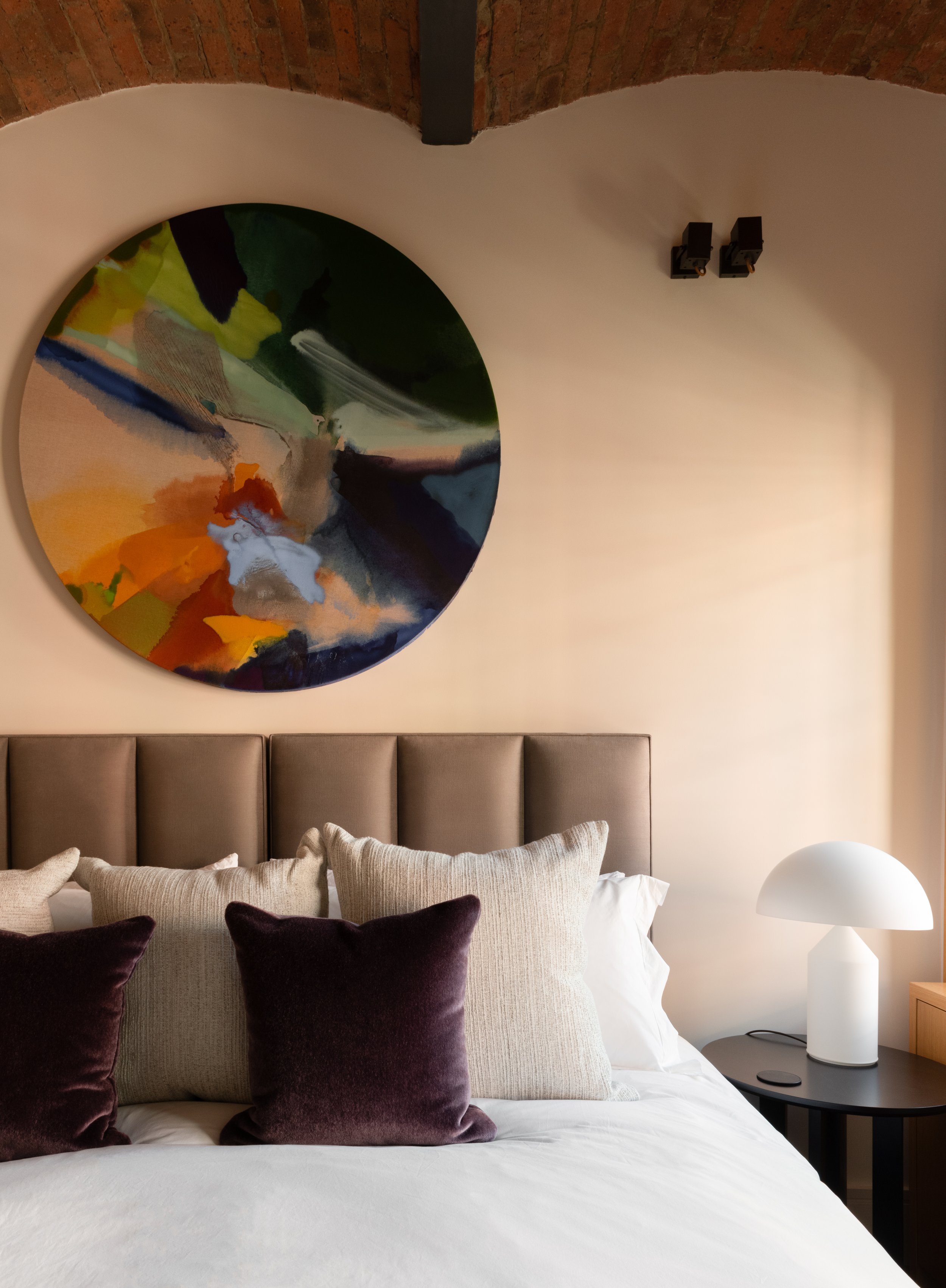 Detail interior photo of a bedroom with a upholstered paneled headboard, and exposed brick ceiling vaults. Above the headboard hangs an abstract colourful round artwork. 