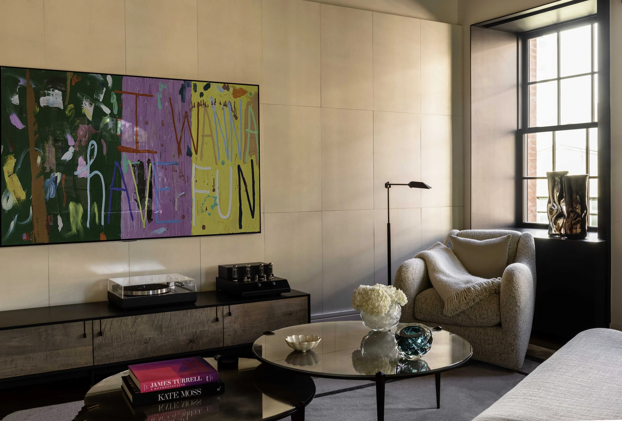 Interior photo of one corner of the sitting room. Light streams in the sash window, lighting the chic but relaxed upholstered furniture.  The wall is covered in a warm neutral toned vertical rectangular panels.