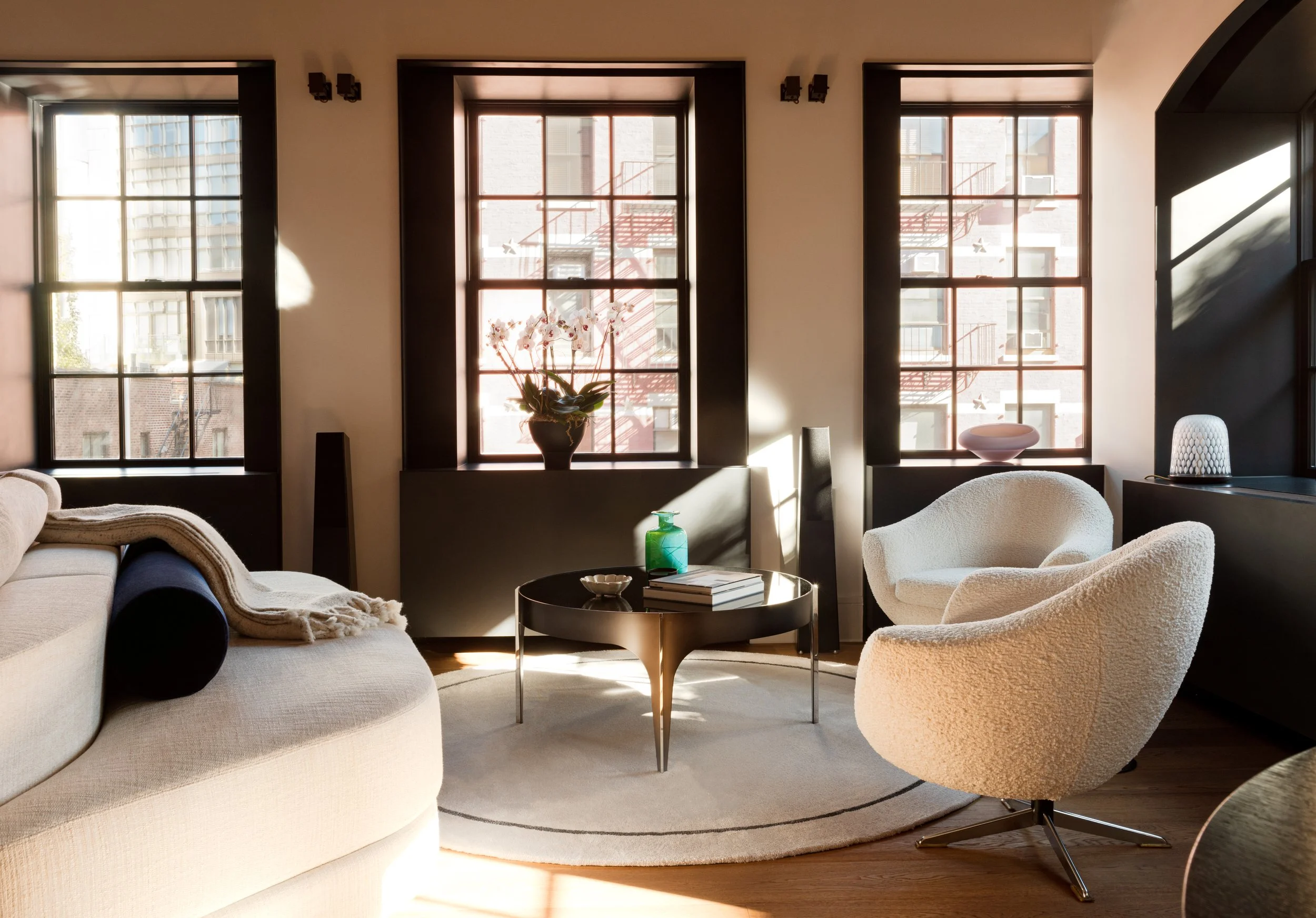 interior photo of a new york city apartment with three windows to one side allowing in light and views of the low-rise brick buildings of NoHo. Inside we have a relaxed sitting area centered on a circular rug. 