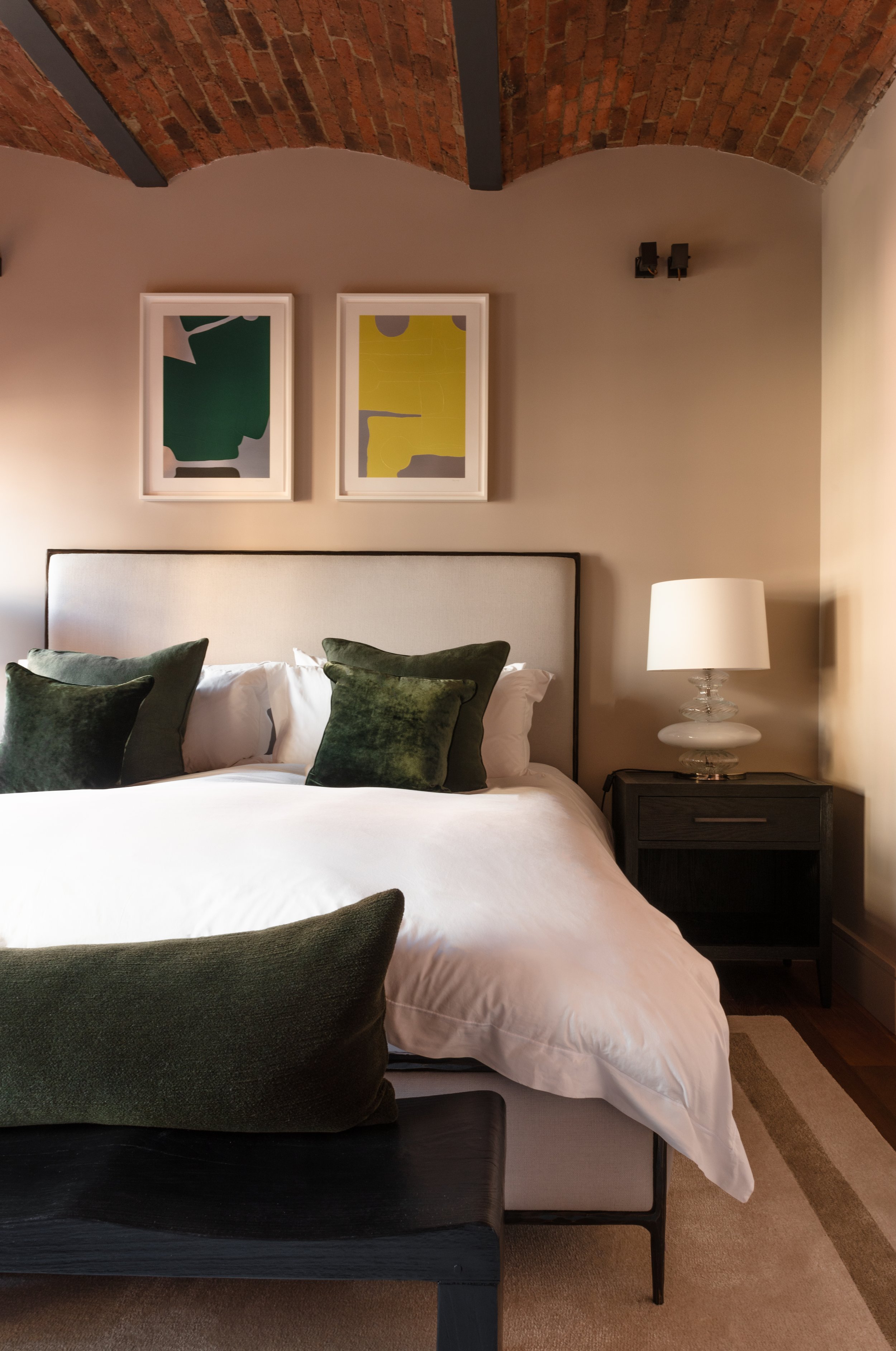 Interior photo of an elegantly appointed bedroom, with a upholstered bed frame, abstract colourful artworks above the bed and exposed brick ceiling vaults. 