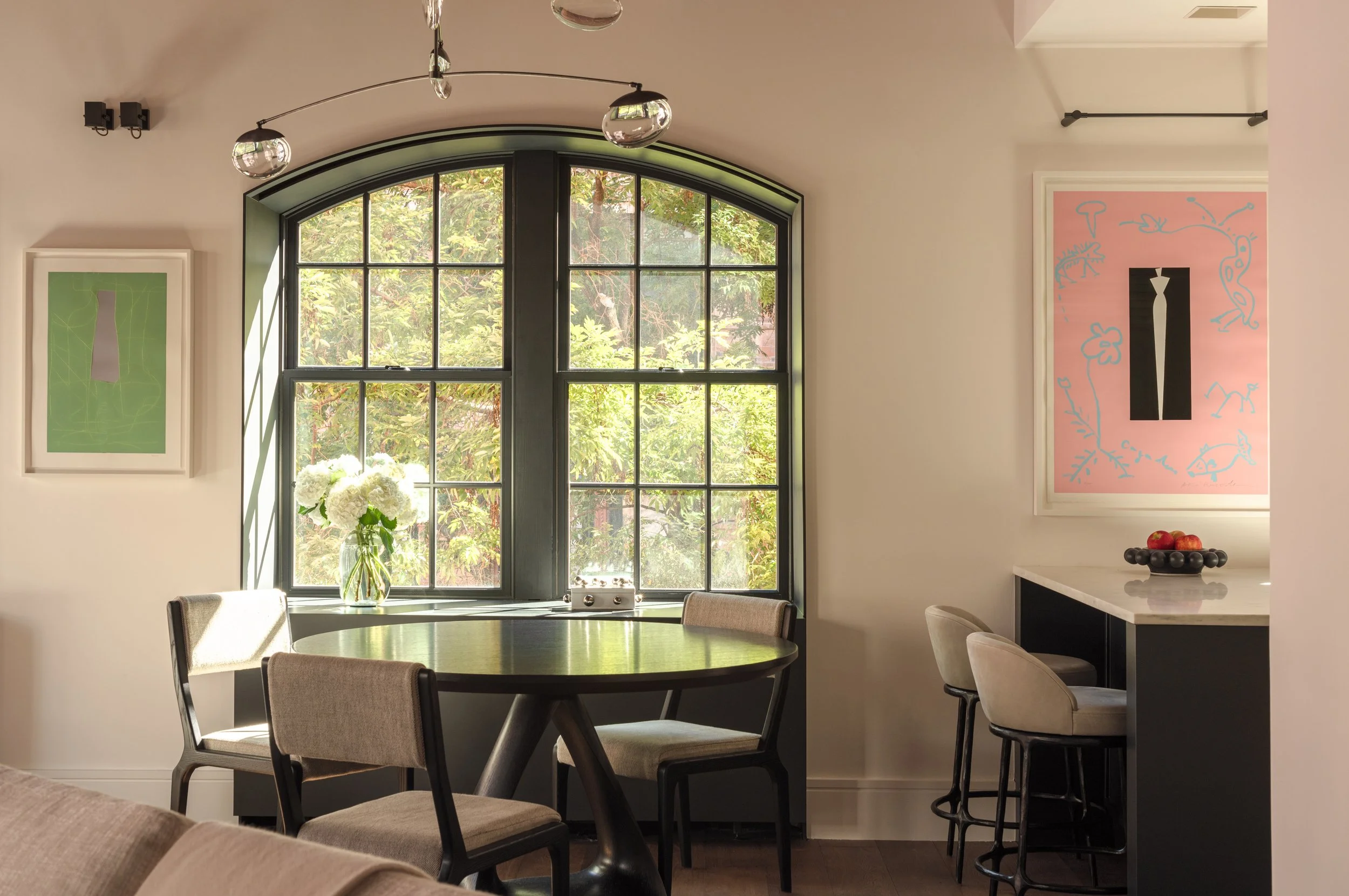 Interior photo of the dining area in front of a large arched window, overlooking a tree-lined street.  To the right is the start of the kitchen, with a pink framed artwork by Aki Kuroda.