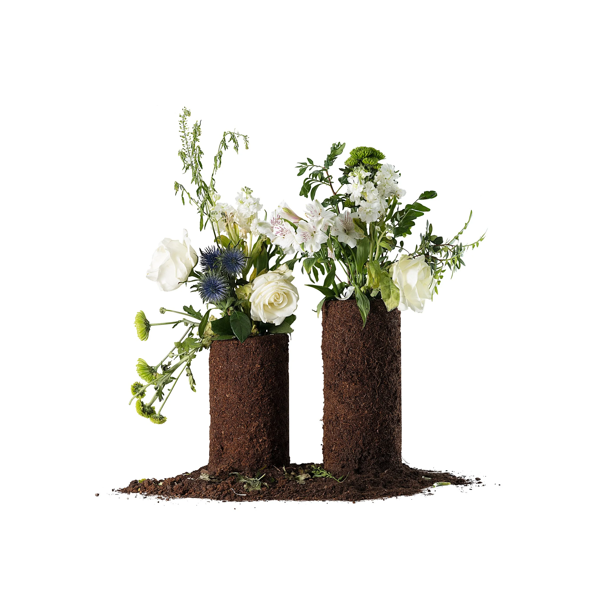 Photo of 2 cylindrical vases constructed of soil, sitting on top of loose soil. Both hold simple rustic bouquets of flowers, in shades of green and white, an artwork by Elaheh Ganji