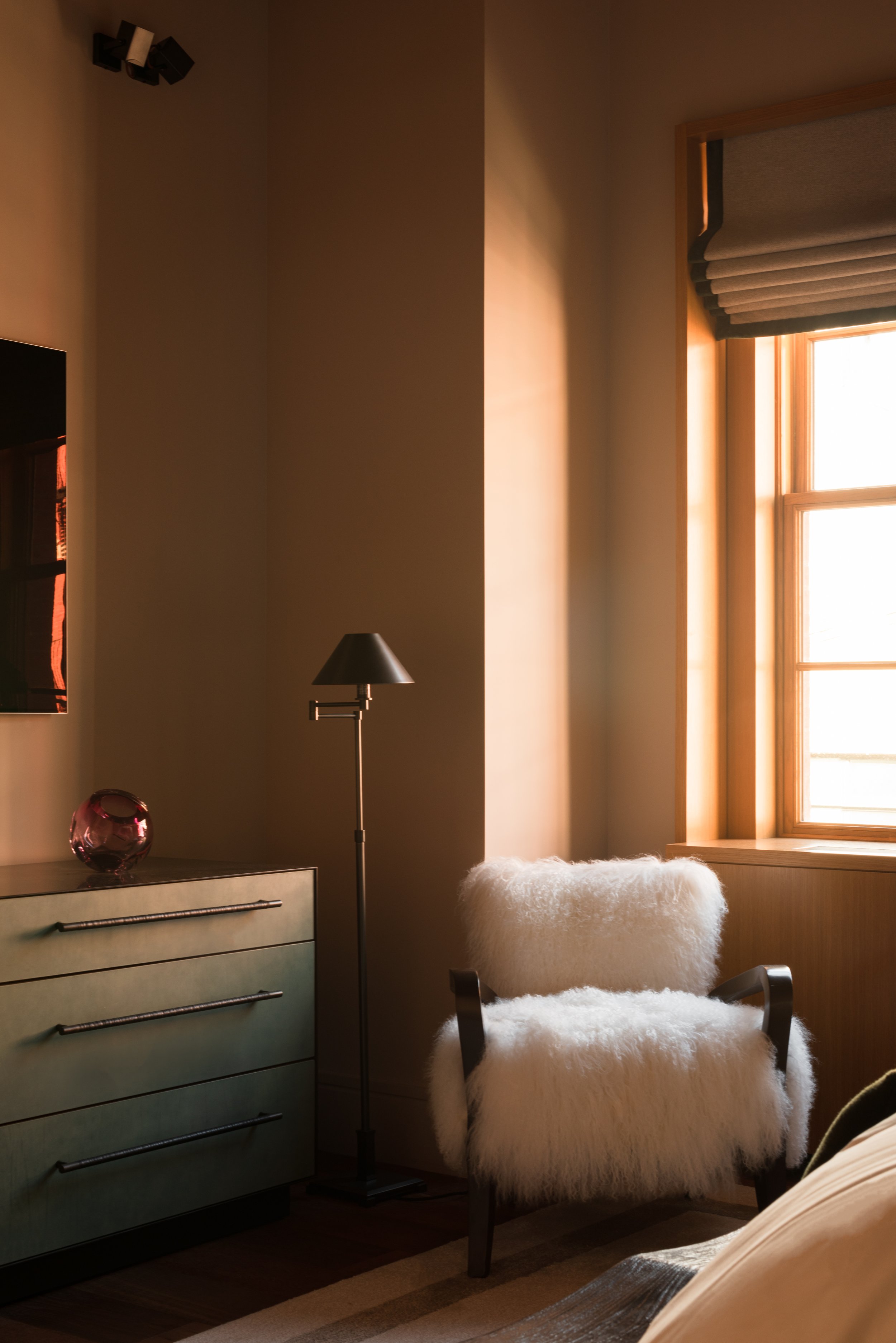 A beautiful moody interior photo of the corner of a bedroom, with shaggy sheep-skin upholstered cafe club armchair by Rabih Hage. Soft light streams in through the sash window. 