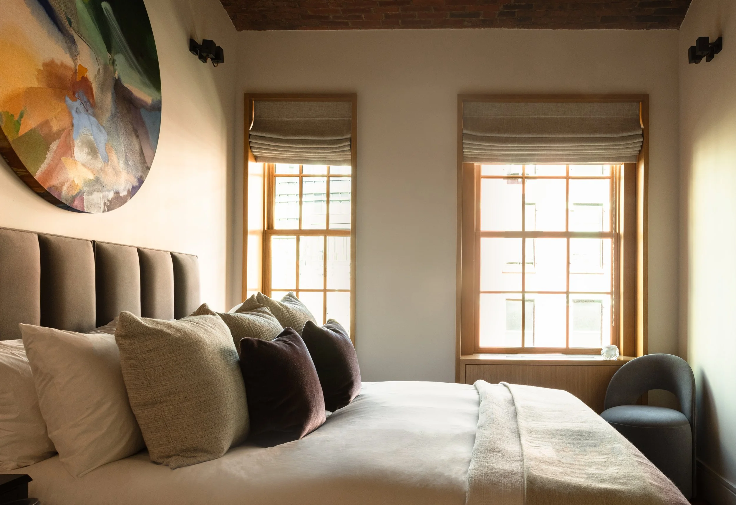 Interior photo of a bedroom, with upholstered headboard, luxurious but understated cushions and bedding, with 2 sash windows dressed with heavy linen roman blinds. Over the bed hangs an abstract colourful round artwork.