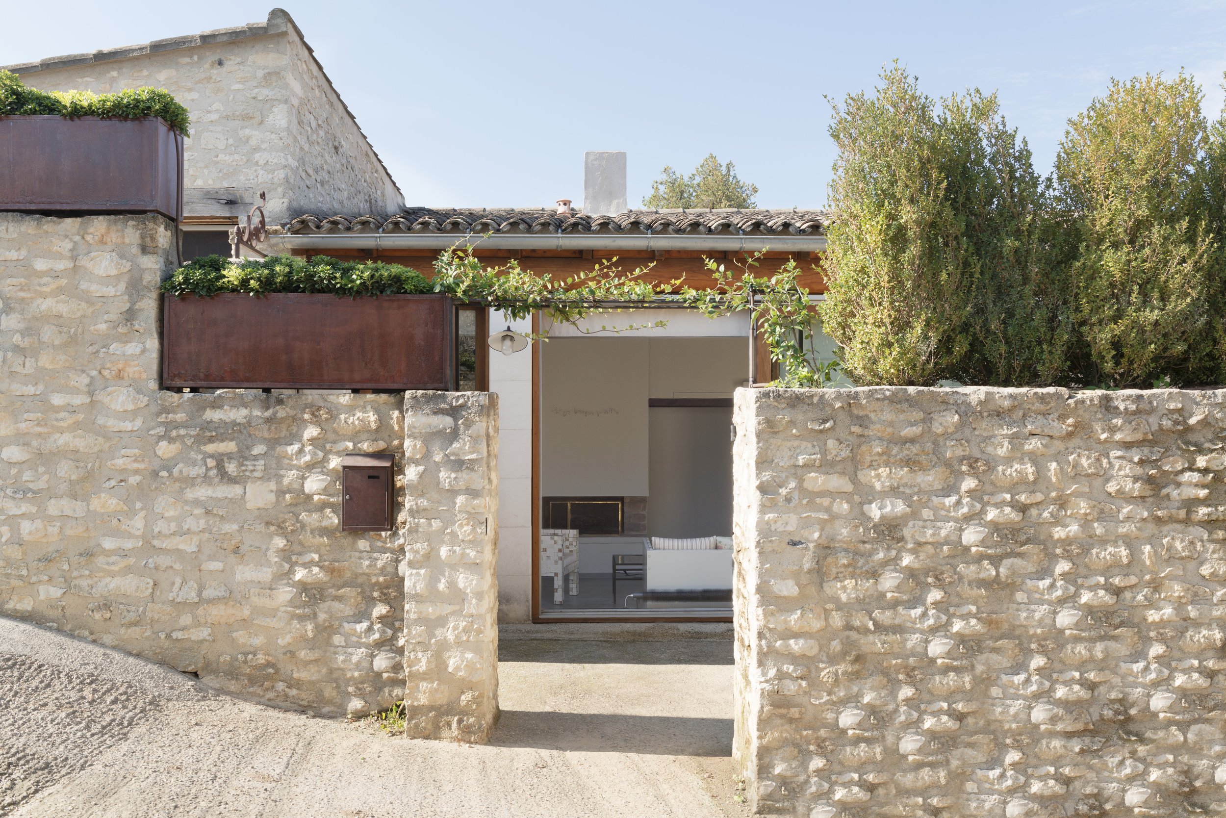 View of the front gateway into the house with stone walls and planters to both sides. Through the gate there is visibility through the floor to ceiling windows to the fireplace inside.