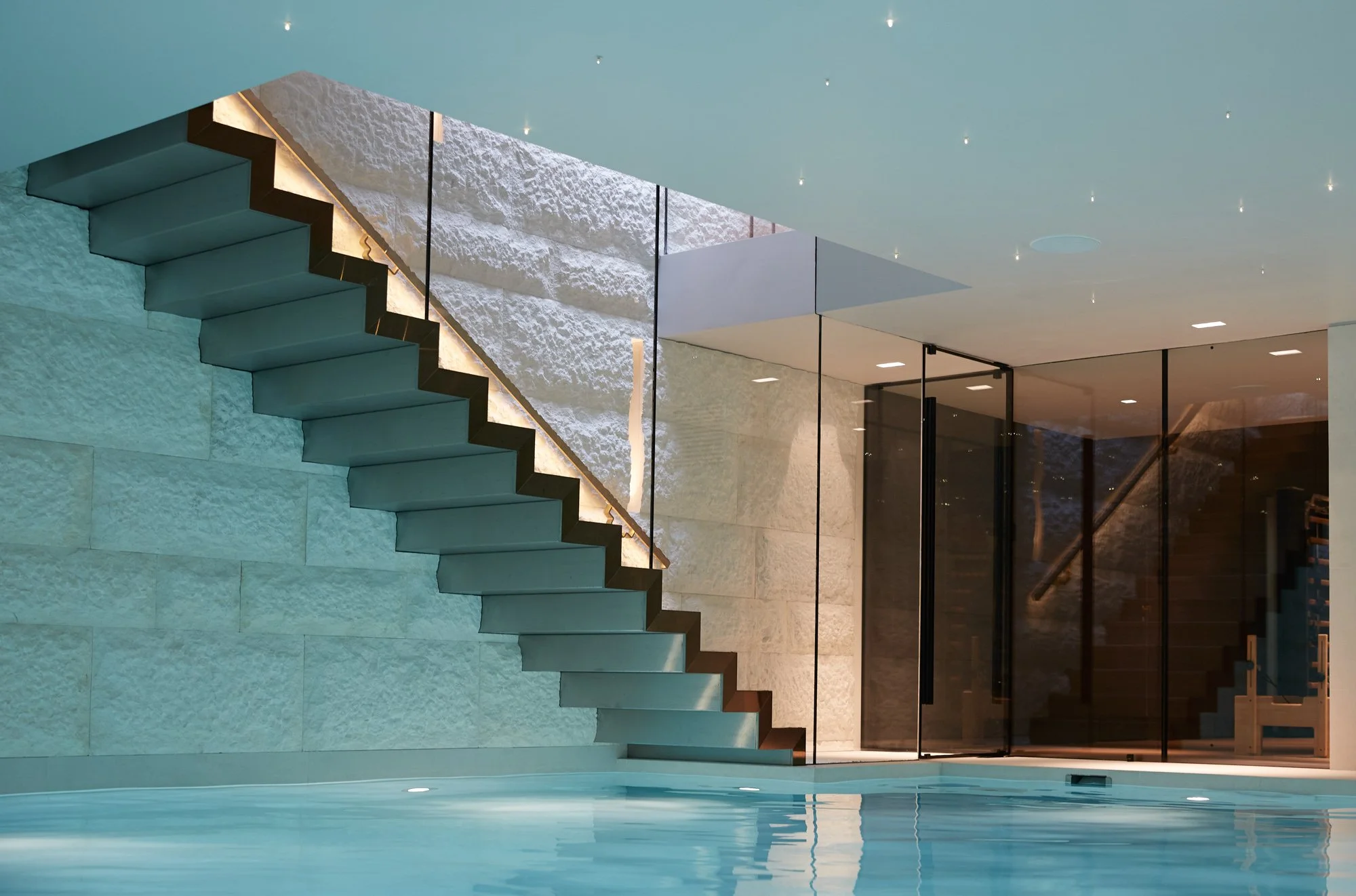 Interior image of a floating staircase projecting from a wall of large textured stone blocks, leading to a basement pool.