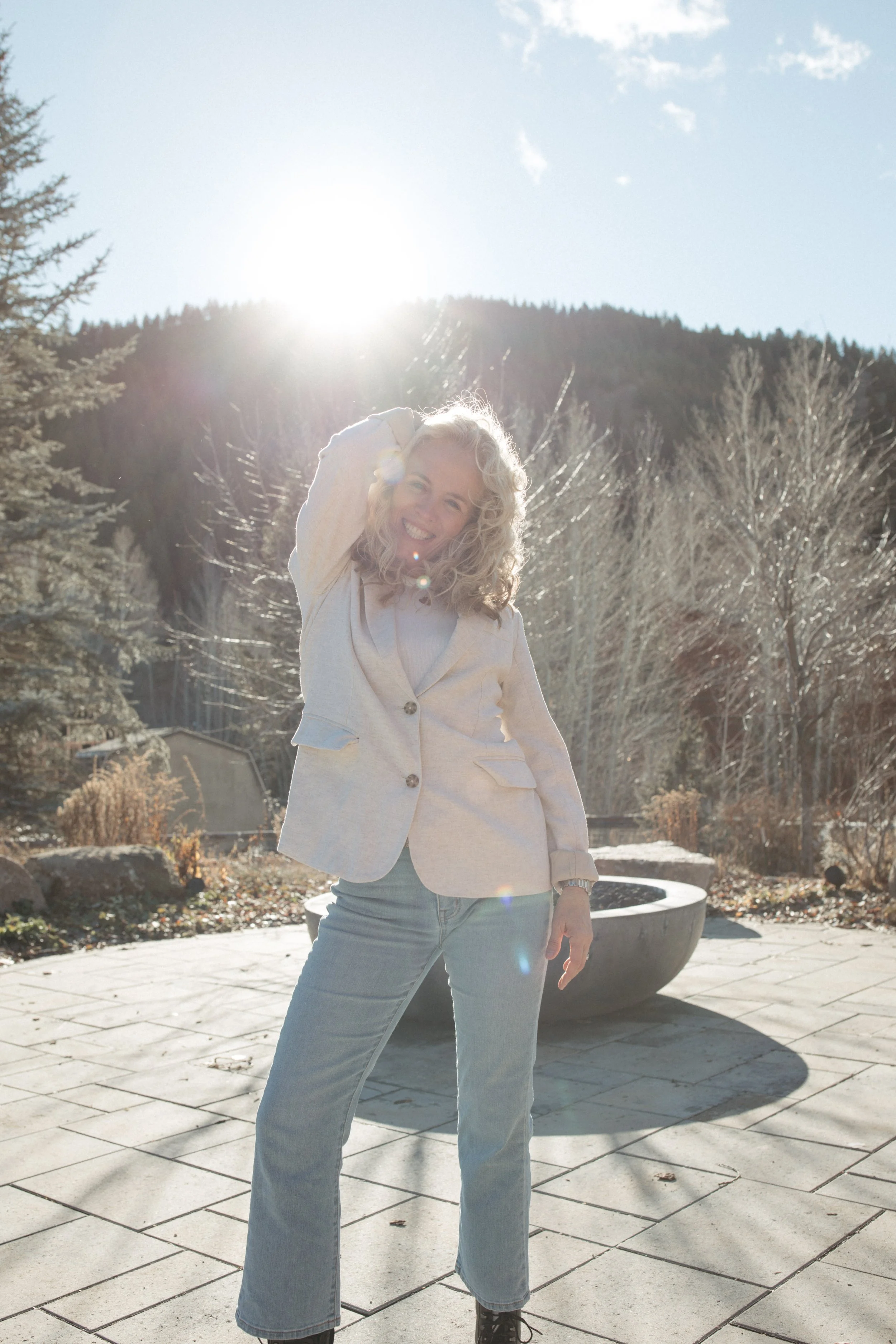 Woman with curly hair smiling outdoors in sunlight, wearing a white blazer and light blue jeans.