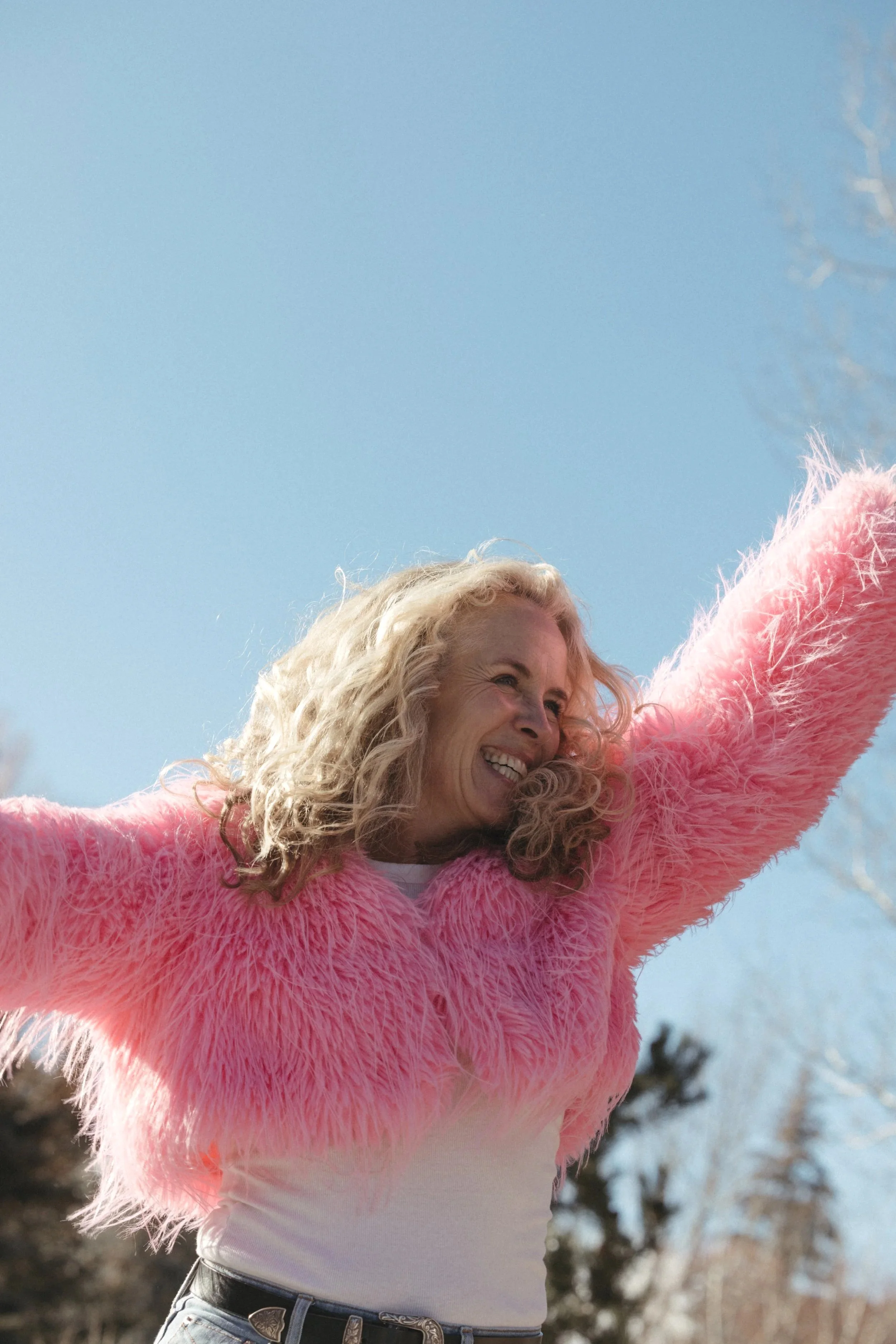 A woman with curly blonde hair wearing a pink fluffy jacket, smiling with arms outstretched against a clear blue sky.