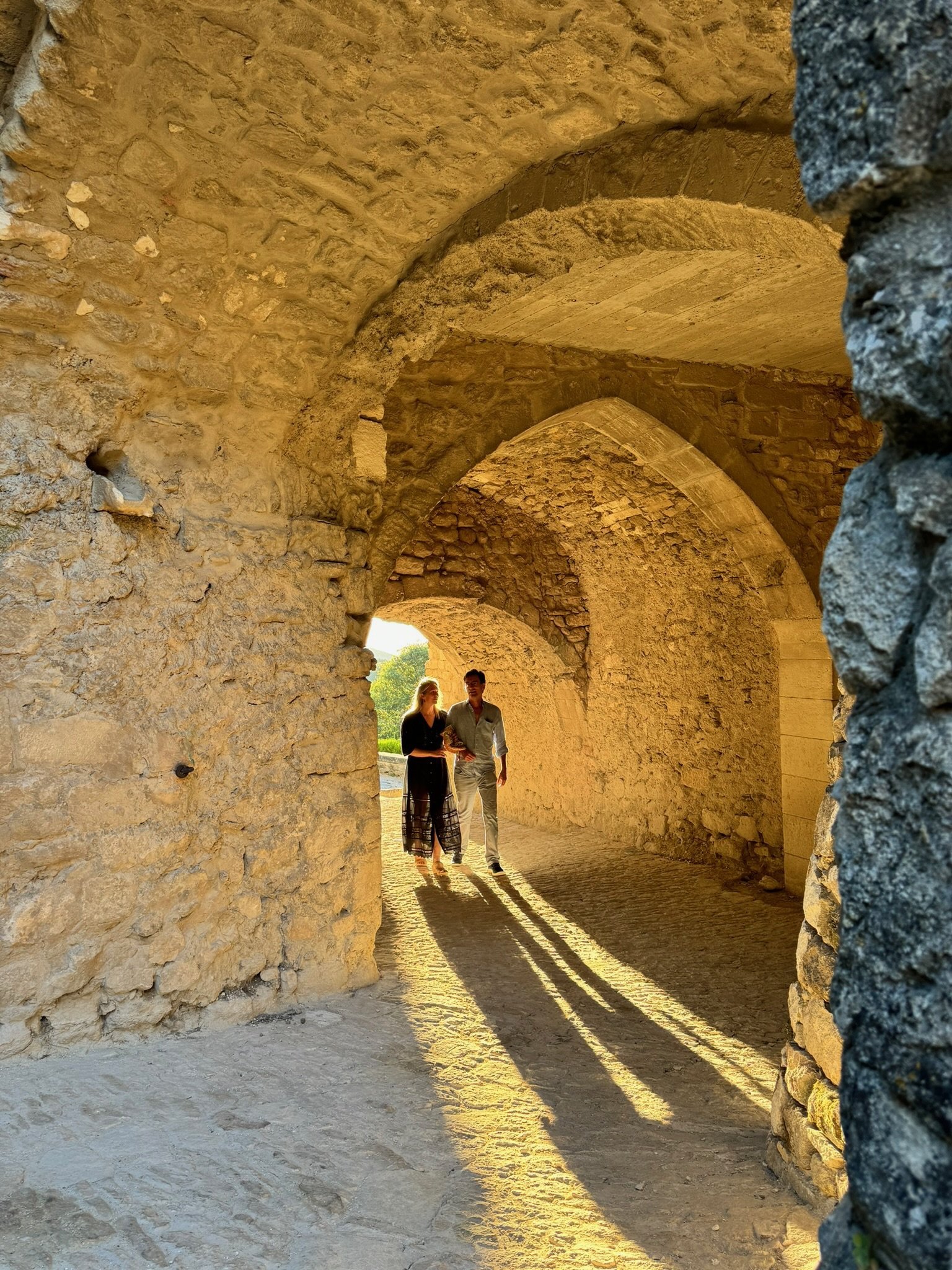 A couple walking hand in hand through an ancient stone archway with warm sunlight casting long shadows behind them.