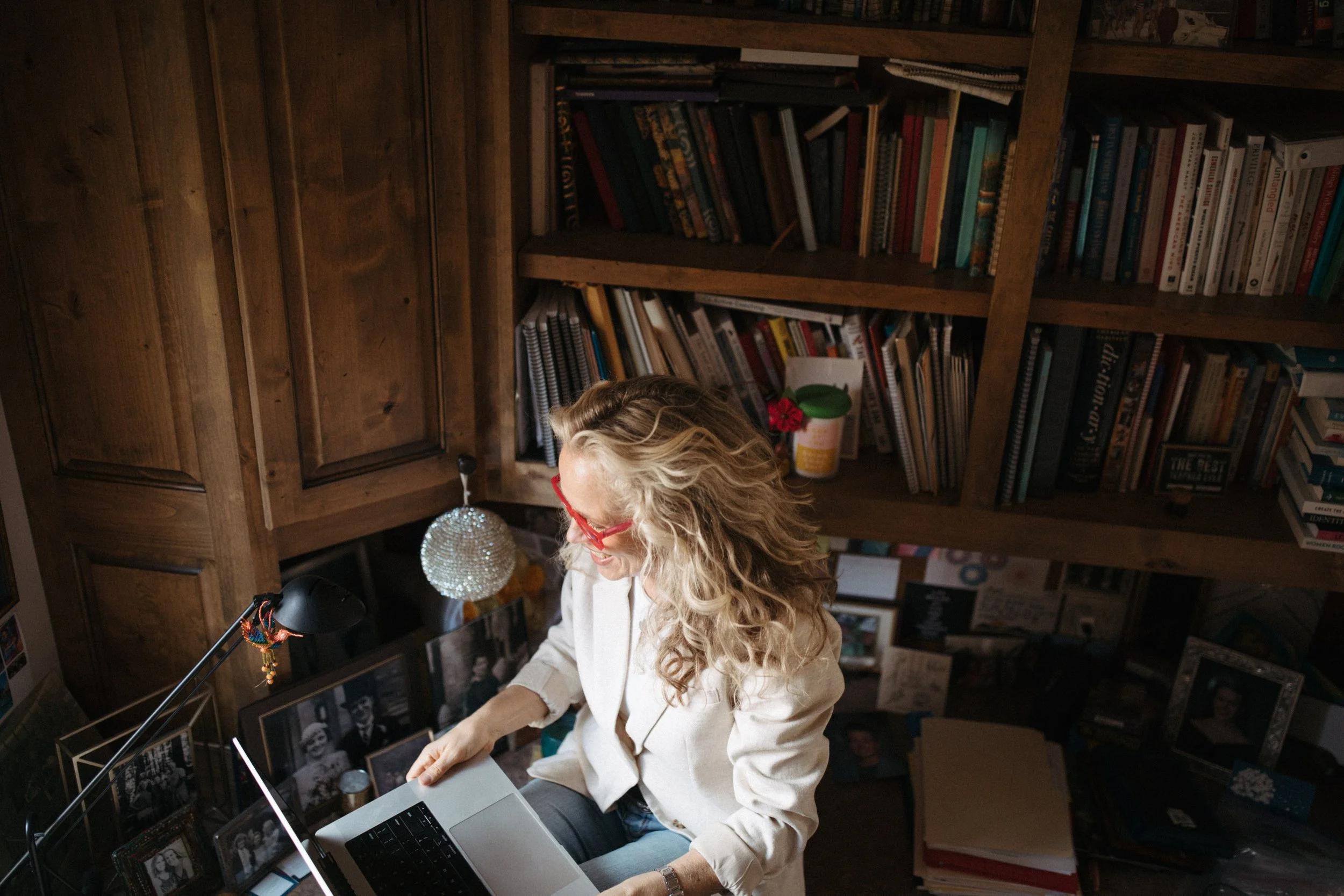 A woman with curly blonde hair, red glasses, and a white blazer working on a laptop in a study with wooden bookshelves filled with books, framed photographs, and miscellaneous items.