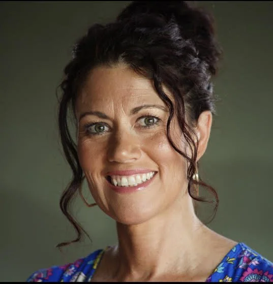 Close-up of a smiling woman with dark, curly hair and blue eyes, wearing a colorful floral top.