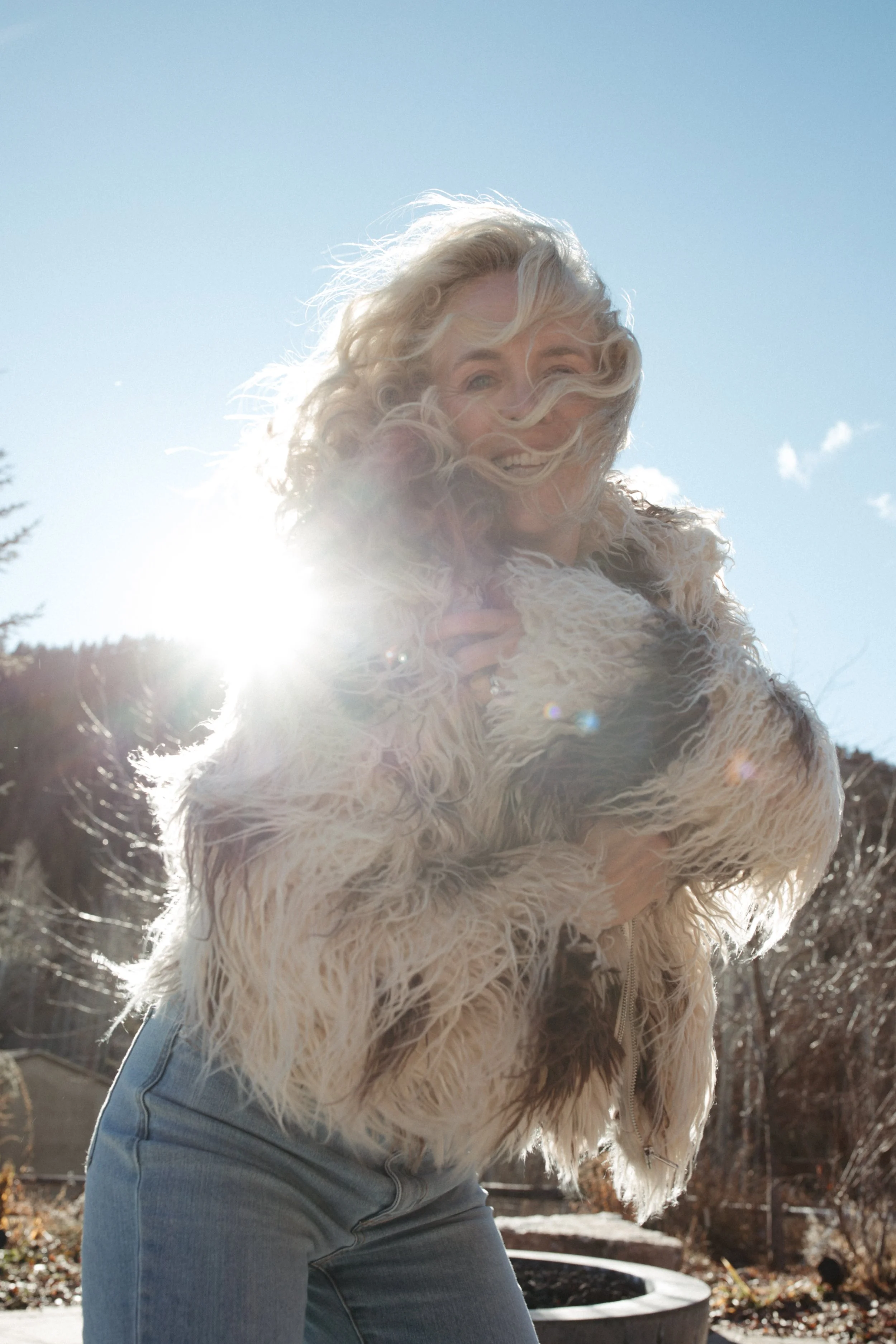 A woman with blonde curly hair wearing a fluffy light-colored jacket and jeans stands outdoors in sunlight, with a faint smile and wind blowing through her hair.