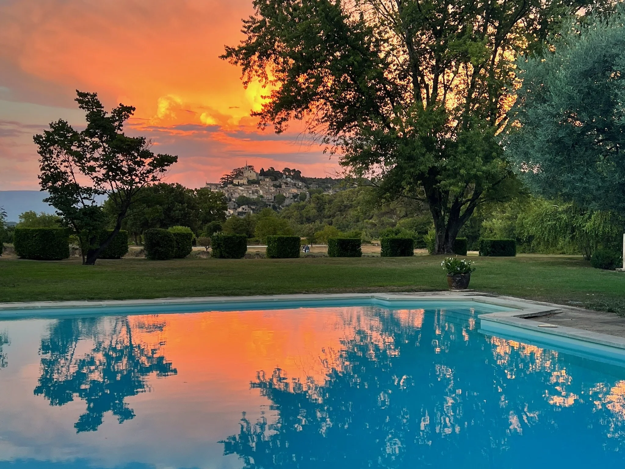 A swimming pool reflecting the colorful sunset sky with hues of orange and pink, surrounded by green trees and well-maintained bushes, with a hillside town in the background.