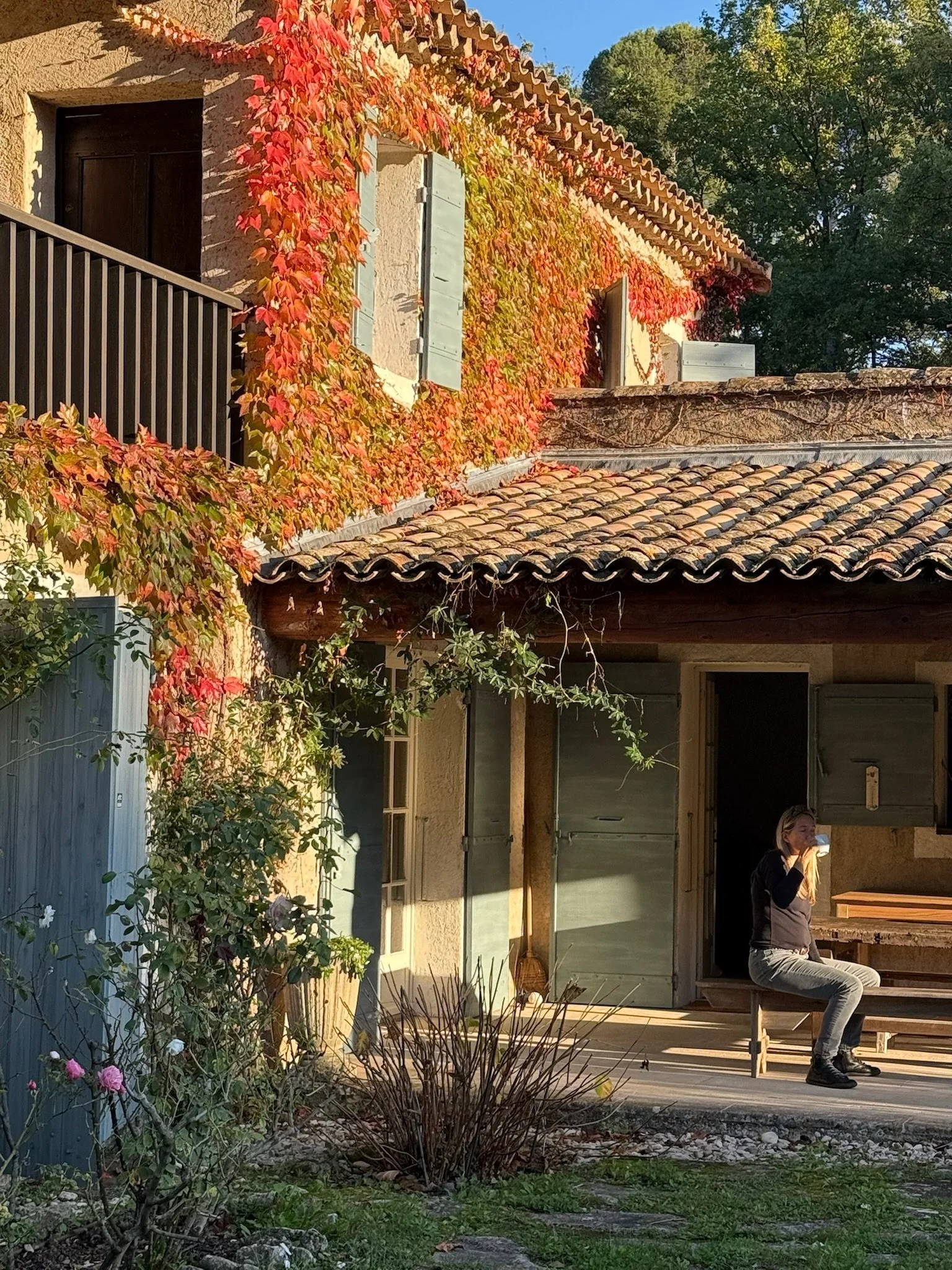 A young woman sitting on a wooden bench eating a meal or snack outside a house with a tiled roof and shuttered windows. The house has colorful autumn leaves growing along the wall, and there are trees in the background.