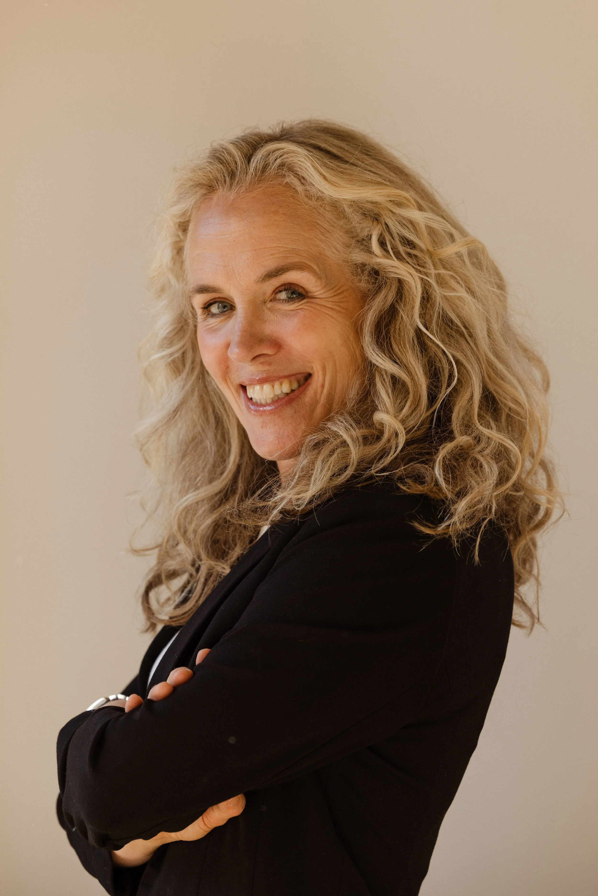 A woman with light skin, blonde curly hair, and blue eyes smiling with arms crossed, wearing a black blazer in front of a neutral background.