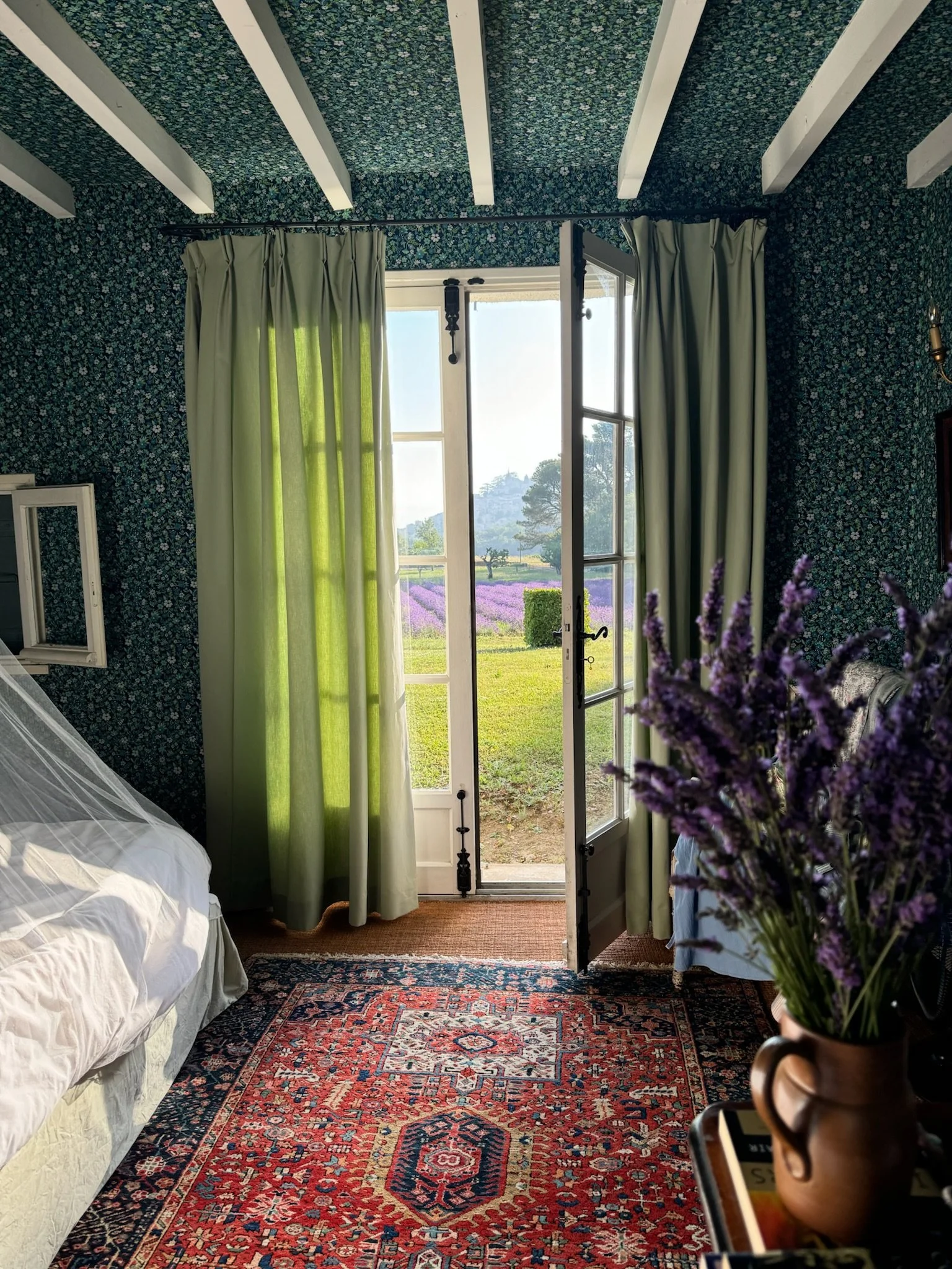 A cozy bedroom with floral wallpaper, green and cream curtains, a view of a lavender field outside the open French door, a patterned area rug, and a vase of purple flowers on a side table.