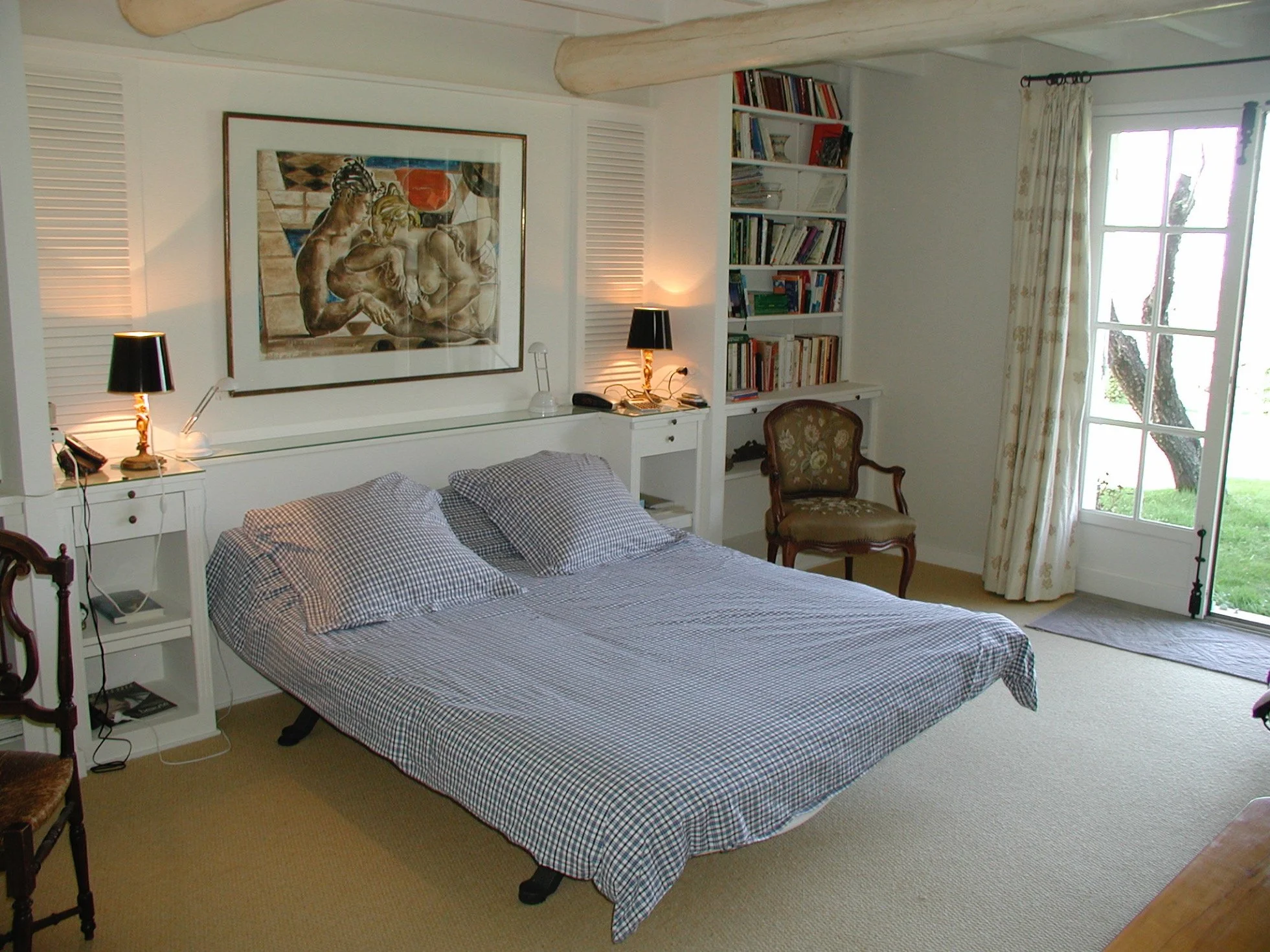 A bedroom with a bed, paintings, lamps, a bookshelf, an armchair, and a glass door opening to outdoors.