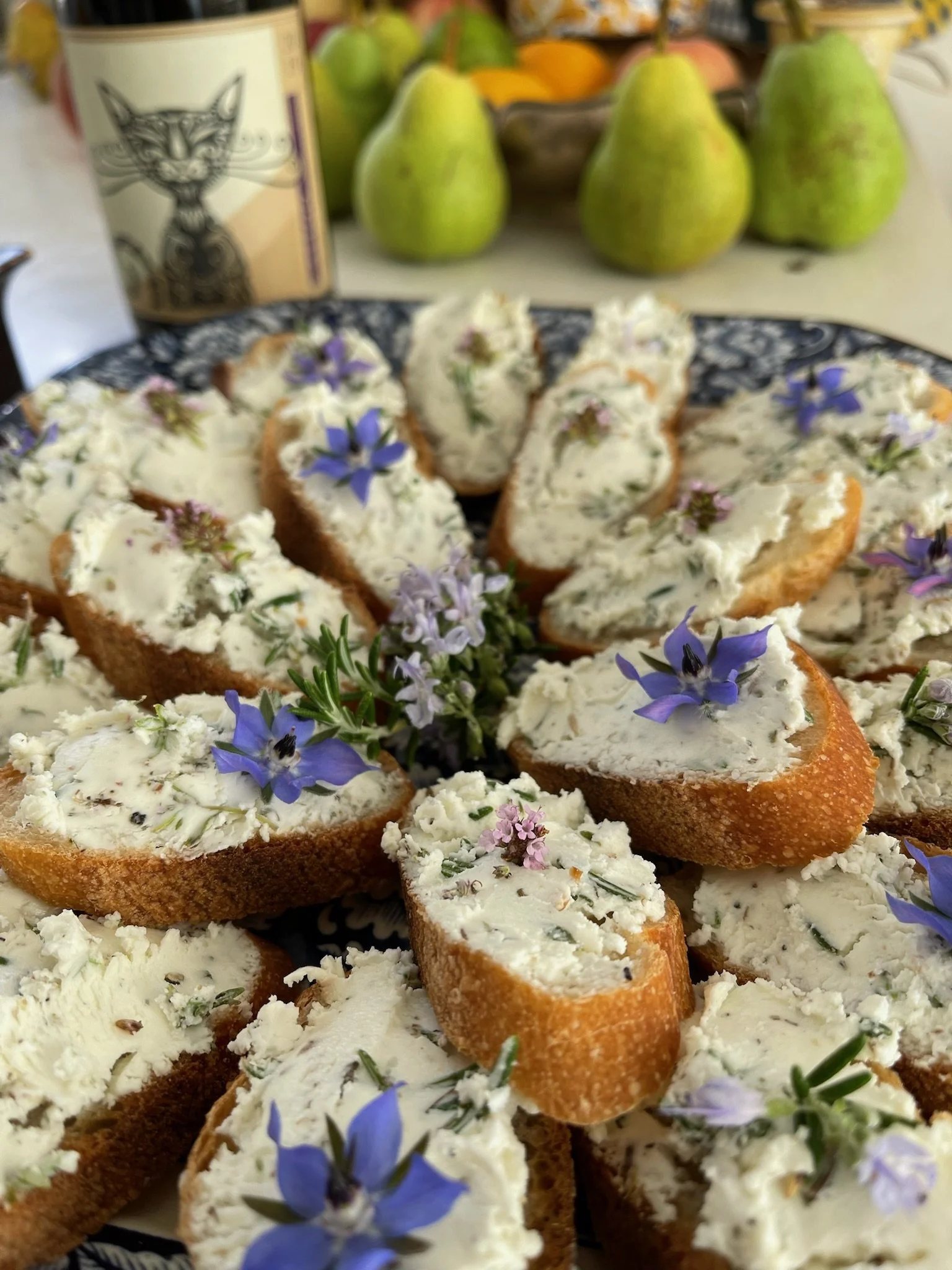 Slices of baguette topped with a creamy herb spread, garnished with purple edible flowers and a sprig of rosemary, arranged on a decorative plate. In the background, whole pears and a bottle with a feline illustration are visible.