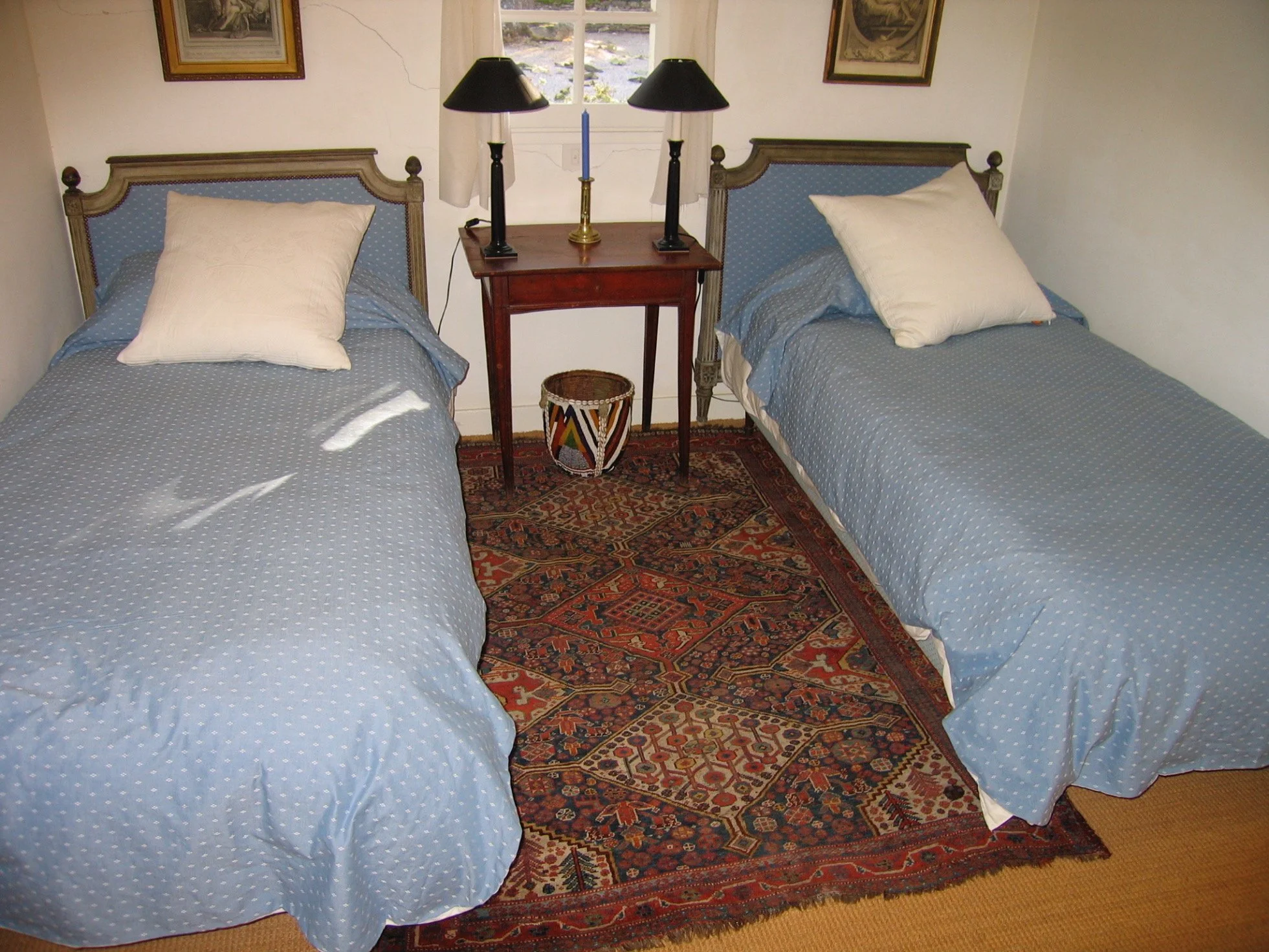 A bedroom with two single beds with blue polka dot bedding, each with a white pillow. Between the beds is a small wooden table with two black lamps, a candle, and a decorative basket underneath. There are framed pictures on the wall and a window behind the table.