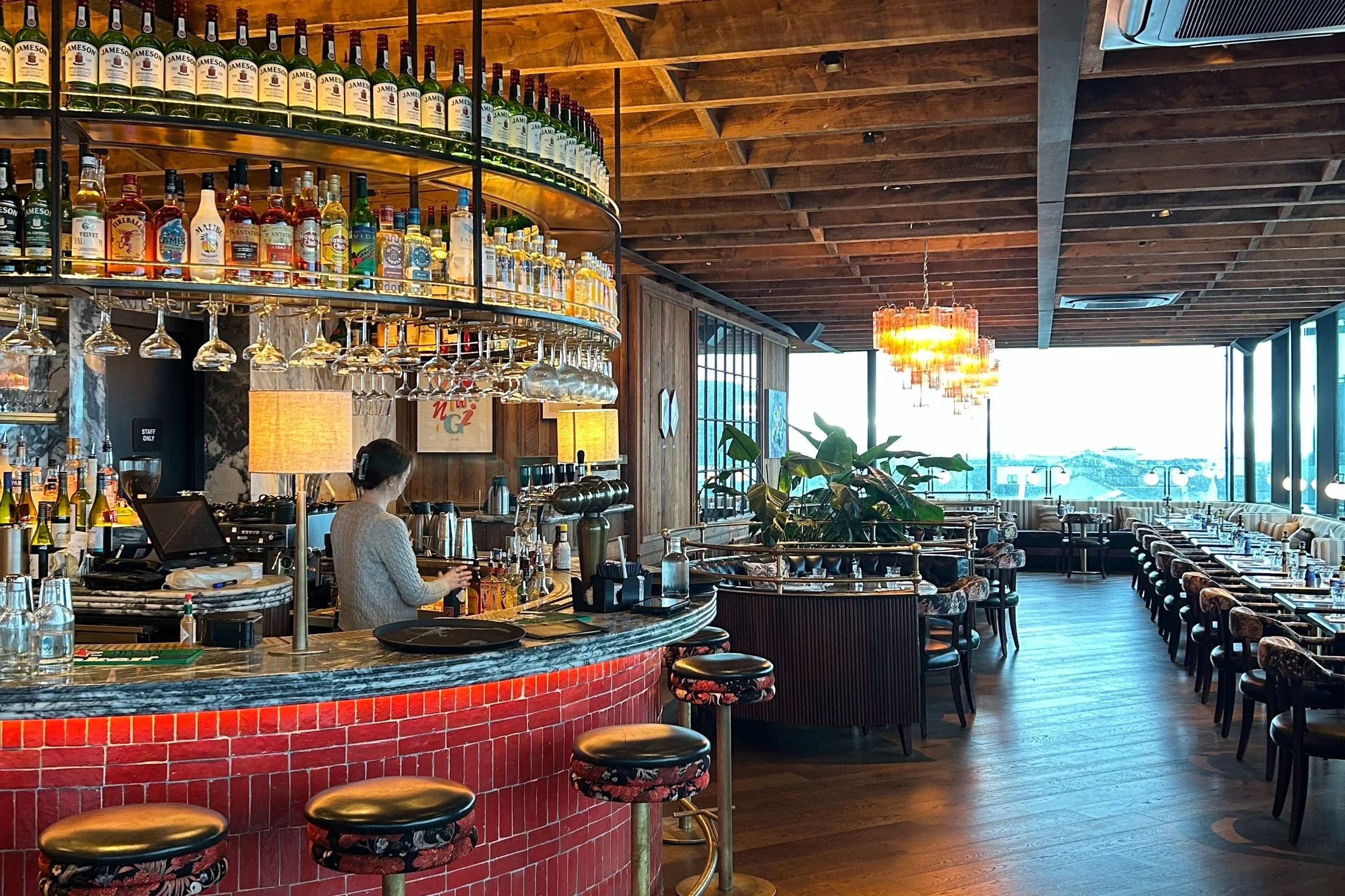 Modern restaurant interior with a bar area displaying various bottles of alcohol and wine glasses hanging upside down. The dining area has tables and chairs arranged near large windows with a view of the outdoors. The space features wooden accents, decorative lighting fixtures, and greenery.