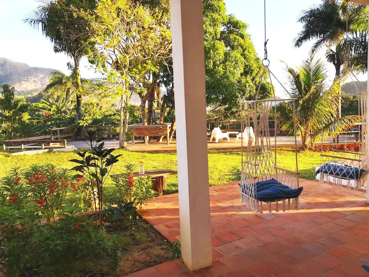 View of a backyard with a brick patio, hanging swing chairs with cushions, lush green lawn, trees, and outdoor seating, with mountains visible in the background under a clear sky.